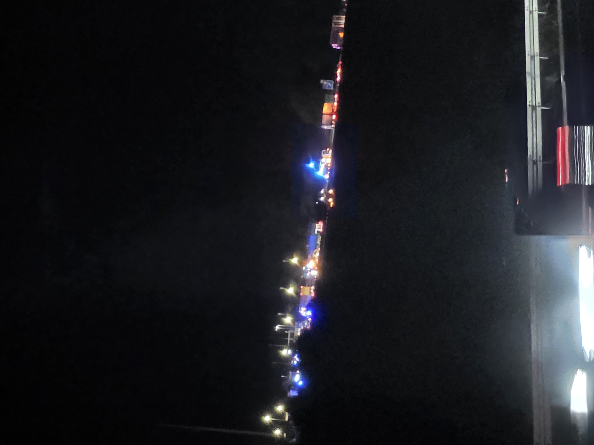 The image depicts a nighttime scene on a highway in Uettingen, Germany. The highway is illuminated by streetlights and the flashing lights of emergency vehicles, suggesting an accident or incident has occurred. Numerous vehicles, including what appear to be police cars with blue flashing lights and possibly ambulances or fire trucks with red and white lights, are visible on the highway. Some of these vehicles are parked, while others may be in motion, indicated by blurred lights. The overall atmosphere is dark and somber, with the emergency lights cutting through the night. The highway itself is a concrete structure with a guardrail, and the surrounding terrain appears to be elevated, possibly a hill or embankment. The foreground of the image shows a portion of the highway barrier, with some light reflections.