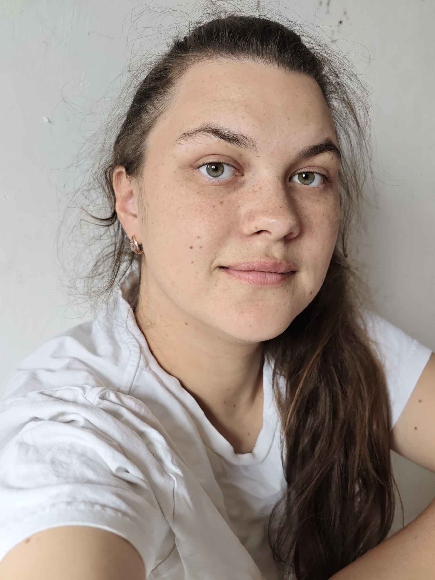 A close-up, headshot-style portrait captures a young adult female looking directly at the camera. She has light skin with prominent freckles across her face, especially on her cheeks, nose, and forehead. Her eyes are green, and her dark brown hair is pulled back, though some fine strands frame her face and escape around her temples. A longer section of her hair falls over her right shoulder. She wears a simple white short-sleeved t-shirt. A small, possibly golden or rose gold, hoop earring is visible in her left ear. Her expression is neutral and calm, with a slight, subtle smile on her lips. The background is a plain, light-colored (off-white or very light grey) wall, which appears to have a slightly uneven or textured surface. The lighting is soft and even, suggesting an indoor setting during the daytime. No text is visible in the image. The location context provided is Noviy, Russia.