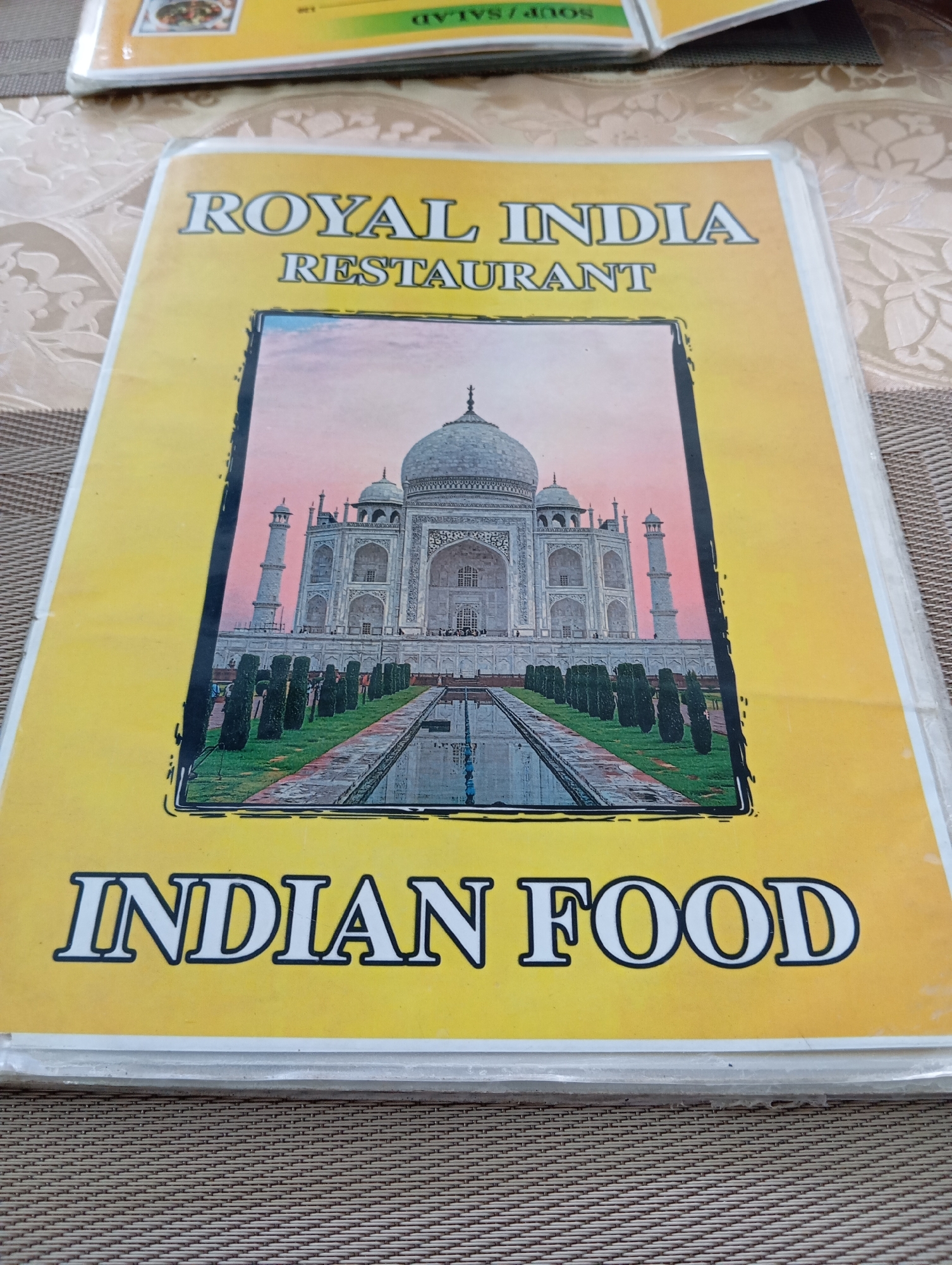 The image shows the cover of a menu for "ROYAL INDIA RESTAURANT INDIAN FOOD". The menu has a yellow background and features a prominent image of the Taj Mahal. The menu is placed on a brown textured surface, possibly a table. In the background, another menu with green and yellow sections is partially visible, with text that reads "SOUP/SALAD". The menu cover appears to be made of laminated paper. The establishment is located in Karon, Thailand.