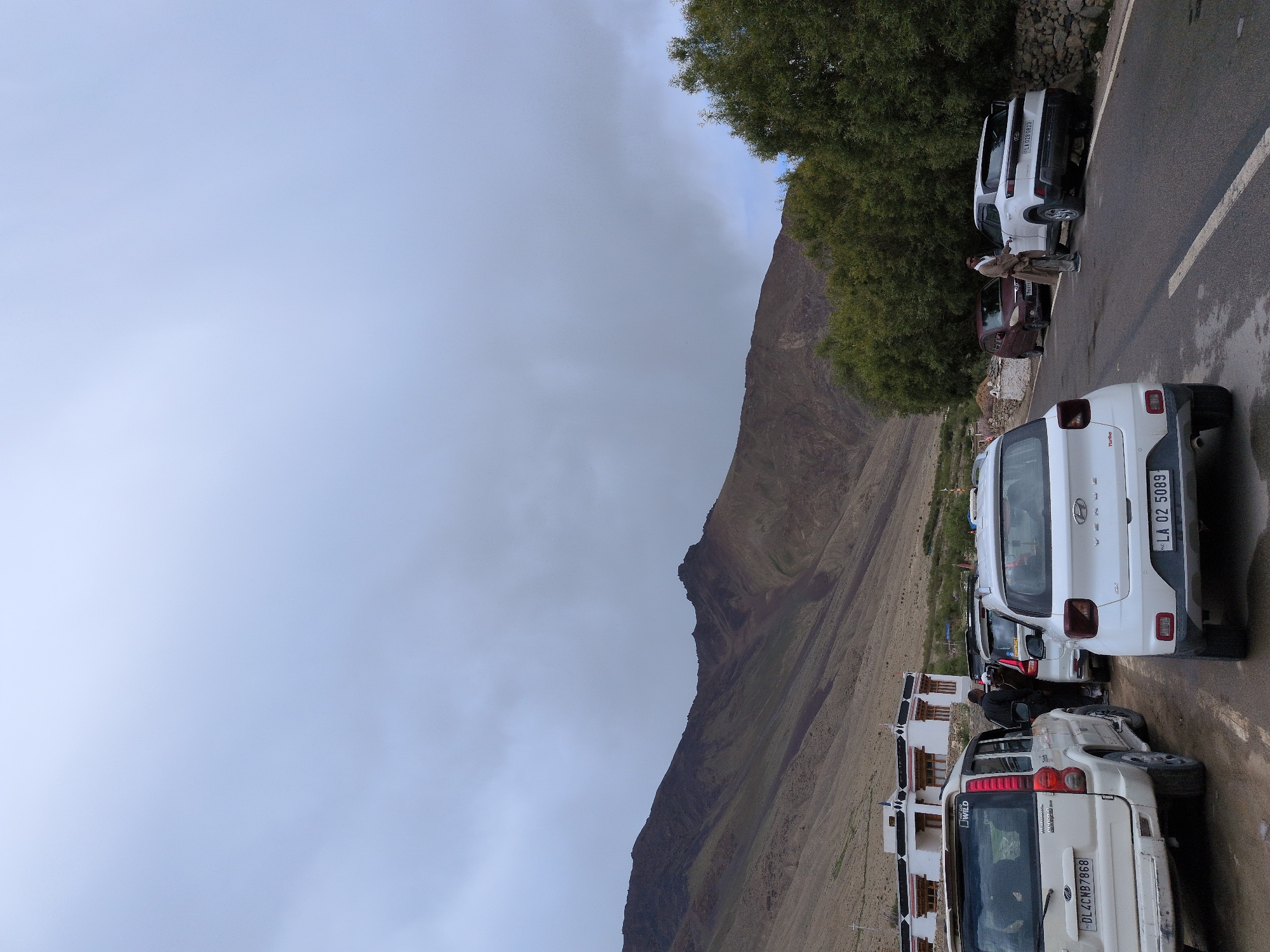 In Khardung, India, under a cloudy sky, a row of parked vehicles lines a paved road. A white Hyundai Venue, license plate "LA 02 5089", is prominent in the foreground, facing right. To its left, a white Mahindra Scorpio, bearing license plate "DL4CNB7868", is parked. Further back, a maroon car and another white SUV are visible. A man in a beige outfit stands near the white SUV, interacting with someone inside. The backdrop features a brown, rocky mountain range and dense green trees. White buildings with red-trimmed windows are situated on the mountainside. The scene appears to be a roadside stop, possibly near a village or a tourist destination.