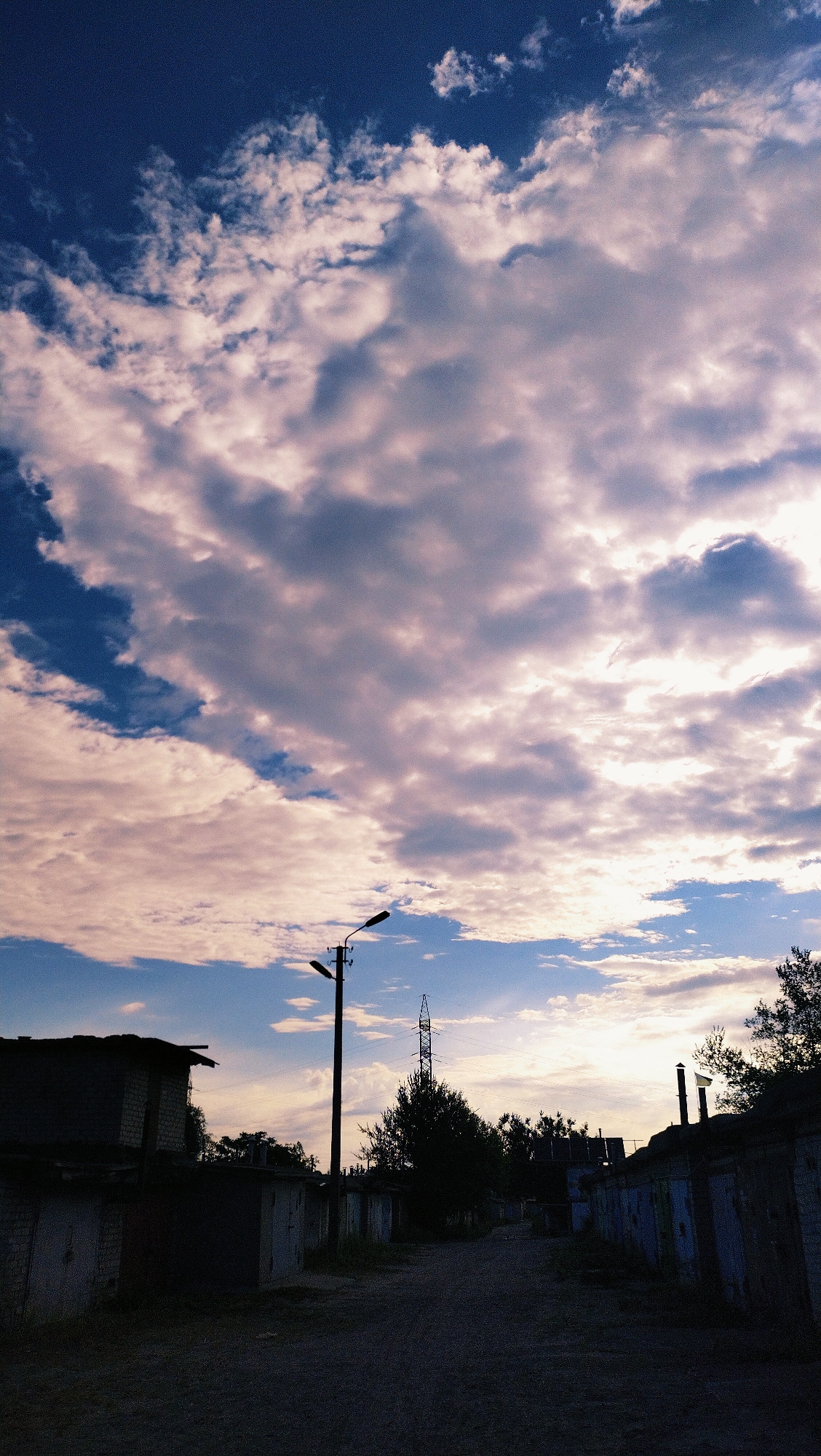 This photograph captures a scene in the city of Horishni Plavni, Ukraine, characterized by a dramatic sky and a row of garages. The sky dominates the upper portion of the image, filled with a multitude of clouds that range from deep blues to soft pinks and whites, illuminated by the setting or rising sun. A single lamppost stands silhouetted against the sky on the left side of the frame, with its light fixture pointing towards the right.
In the foreground and mid-ground, a collection of garages is visible, stretching along a dirt road. These structures appear weathered and somewhat dilapidated, with varied door designs and visible brickwork. Some of the garages are in deep shadow, emphasizing the contrast with the brightly lit sky. Trees are sparsely scattered behind the garages, and a tall transmission tower is visible in the distance, further adding to the urban industrial landscape. The overall atmosphere is one of quietude and transition, perhaps during dusk or dawn, with the natural beauty of the sky juxtaposed against the man-made structures. There are no people or animals visible in the image.