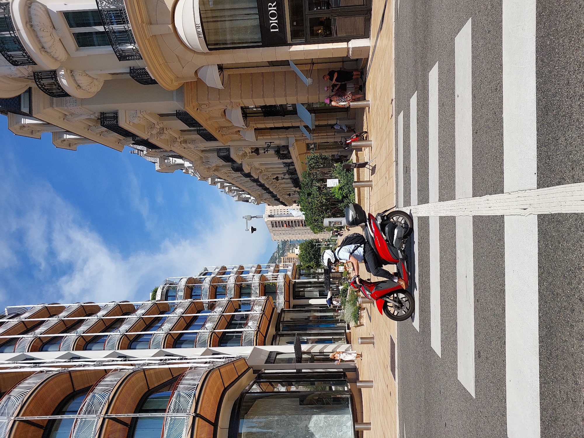 The image shows a man riding a red scooter across a crosswalk on a sunny day in Monaco. The man is wearing a white helmet, a light blue shirt, and dark pants, and has a black backpack. To the left of the scooter is a modern building with curved balconies and large windows. To the right is a more traditional building with ornate details, balconies, and awnings, including a prominent "DIOR" sign. Several people are visible on the sidewalk in front of the Dior store. In the background, other buildings and hills can be seen. The street is paved with asphalt, and the crosswalk is marked with white stripes. The sky is blue with some white clouds.