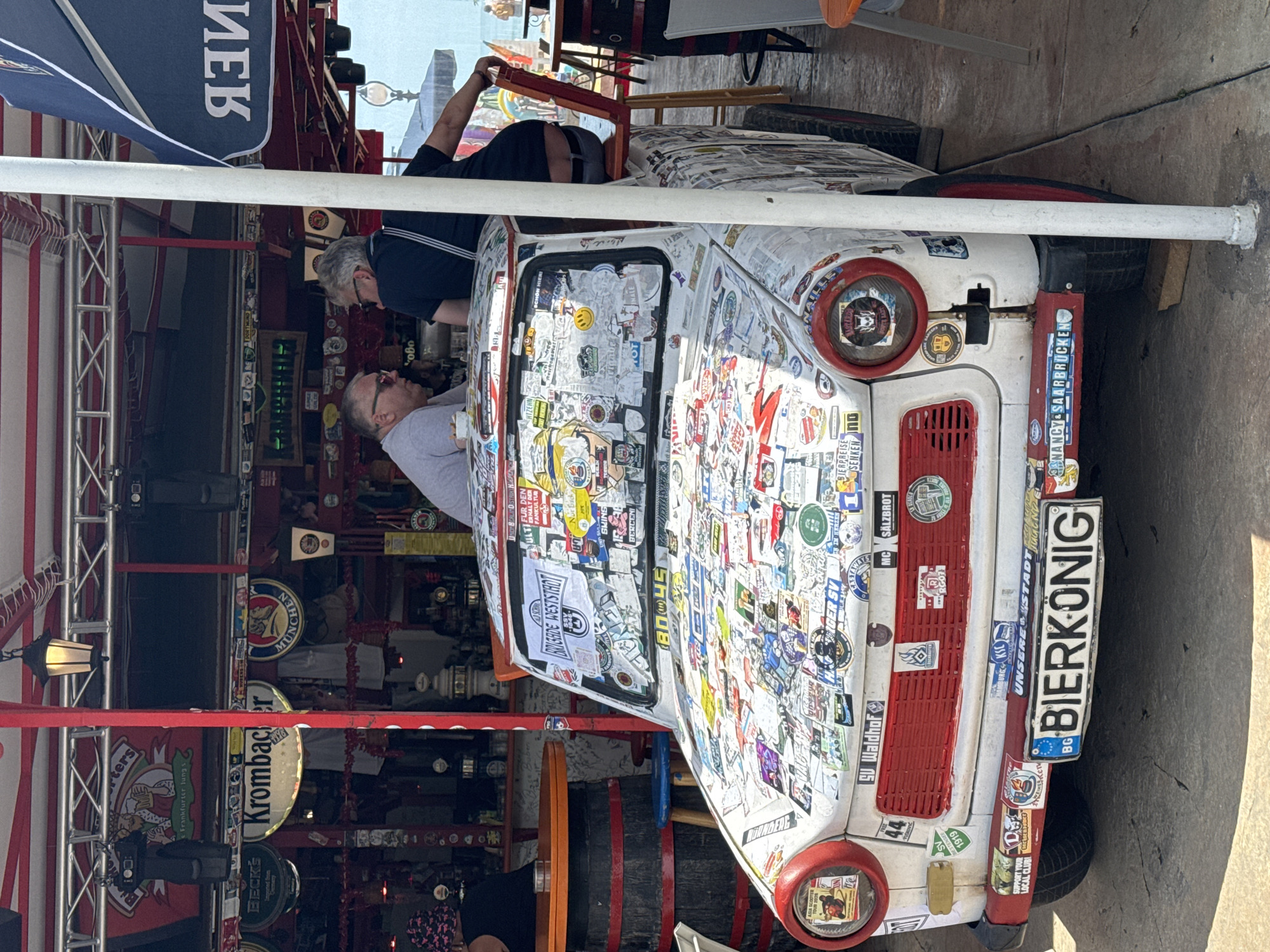 The image captures a white Trabant car, heavily adorned with numerous stickers, parked outdoors in what appears to be a casual outdoor establishment. Two men are seated near the car. One man, wearing sunglasses and a light-colored shirt, is looking towards the car. The other man, wearing a dark shirt, is seated on a red chair with his back partially to the camera. The car is positioned under a red and white canopy structure, with exposed metal beams and lighting fixtures. Behind the car and canopy, a bar area is visible with bottles and what appear to be beer taps. The environment suggests a place that might be a bar or restaurant with an outdoor seating area. The presence of a license plate with "BIERKONIG" and Bulgarian license plate characters suggests the car is registered in Bulgaria, and the overall ambiance, including the casual attire of the people and the outdoor setting, points to a leisure or entertainment venue. Specifically, the establishment is located on the beach alley in Varna, Bulgaria, as evidenced by the background elements and the general setting.