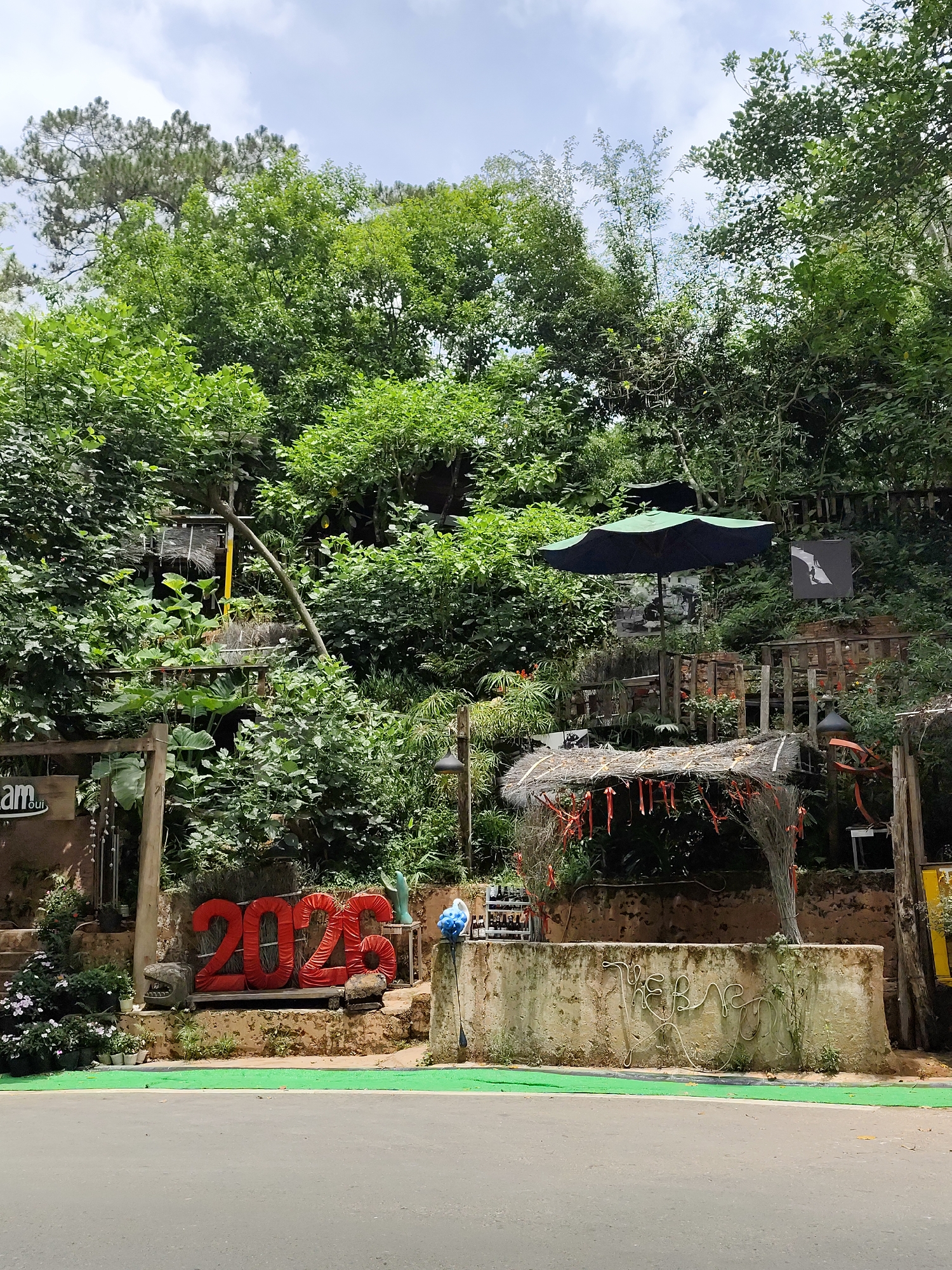This image depicts a view in the city of Da Lat, Vietnam, with a street in the foreground. Behind the street is a low concrete wall, decorated with a rope design. Behind the wall is an elevated area overgrown with plants and trees. Red numbers spelling "2023" are displayed on the elevated area behind the wall. Several decorative elements are visible among the plants, including a thatched structure adorned with red ribbons, a dark green umbrella, and wooden structures.