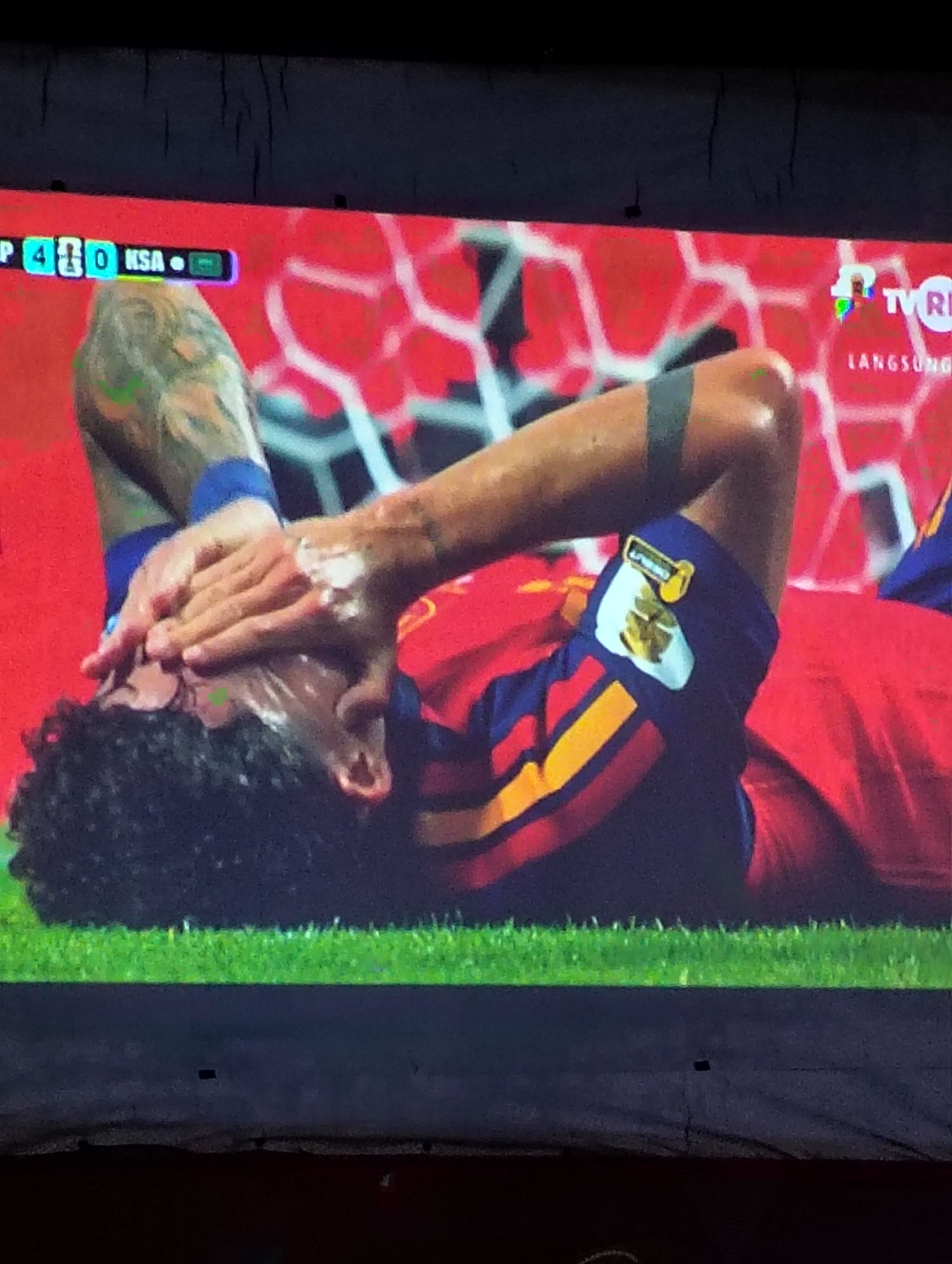 The image shows a close-up view of a male soccer player lying on a grassy field with his hands covering his face. He is wearing a red and blue jersey with yellow accents and a black armband. The score "480" is visible in the upper left corner of the screen, along with "KSA" and a flag. In the upper right corner, a TV logo and the word "LANGSUNG" (which means "live" in Indonesian) are displayed. The background appears to be a stadium with red netting. The scene is not specifically located in the city of Payagaboh, Indonesia; it is a broadcast of a soccer match.