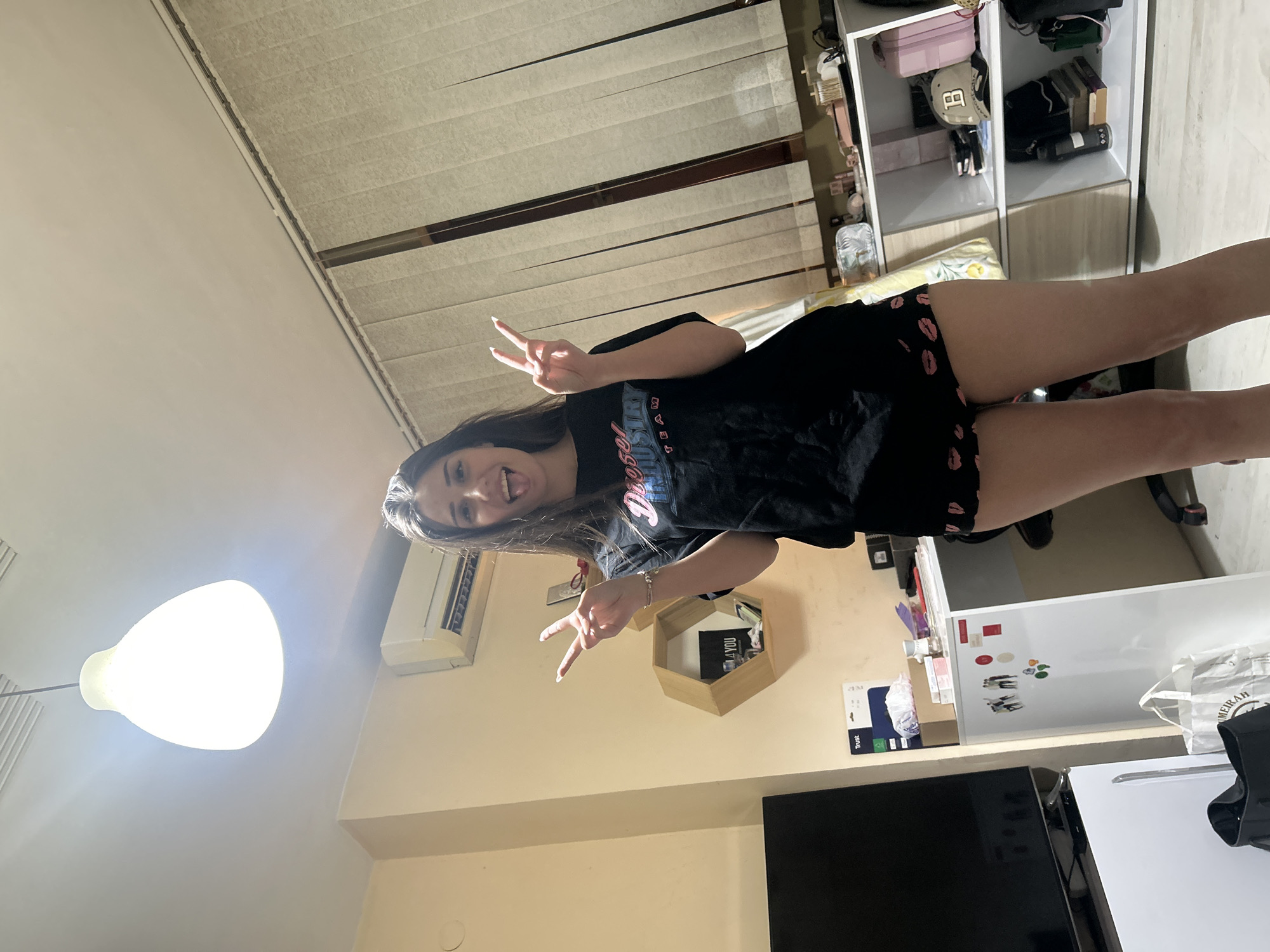An indoor scene depicts a young female standing center-frame, illuminated by a prominent ceiling light. The female has long, dark brown hair and is smiling broadly with her tongue extended, making a peace sign (V-gesture) with both hands. She is wearing a black short-sleeved t-shirt with the text "Diesel Industry Team" printed on the front, and black shorts adorned with pink lipstick kiss marks.
To the left, a large, dark flat-screen television is visible, with a white storage unit or mini-fridge positioned beneath it. A white mini-fridge, decorated with several magnets, stands next to the TV. A "Trust" brand product box and other items rest on a surface behind the fridge. A white bag and a black bag are on the floor in the lower left corner.
On the right side of the frame, a window is covered by light-colored vertical blinds. A white desk or low cabinet extends from the center-right towards the right, holding various small objects. To its right, a white shelving unit contains items such as a pink case, a beige baseball cap with a dark "B" logo, a black water bottle, and a stack of books. A black office chair is partially visible behind the female's legs.
The room features light-colored walls, with a white wall-mounted air conditioning unit located on the upper left wall. A wooden hexagonal wall shelf, holding a dark object with white text, is mounted on the wall behind the female. The ceiling is white, and the floor appears to be a light-colored hard surface. No discernible elements in the image provide specific information to confirm the location as Plovdiv, Bulgaria.