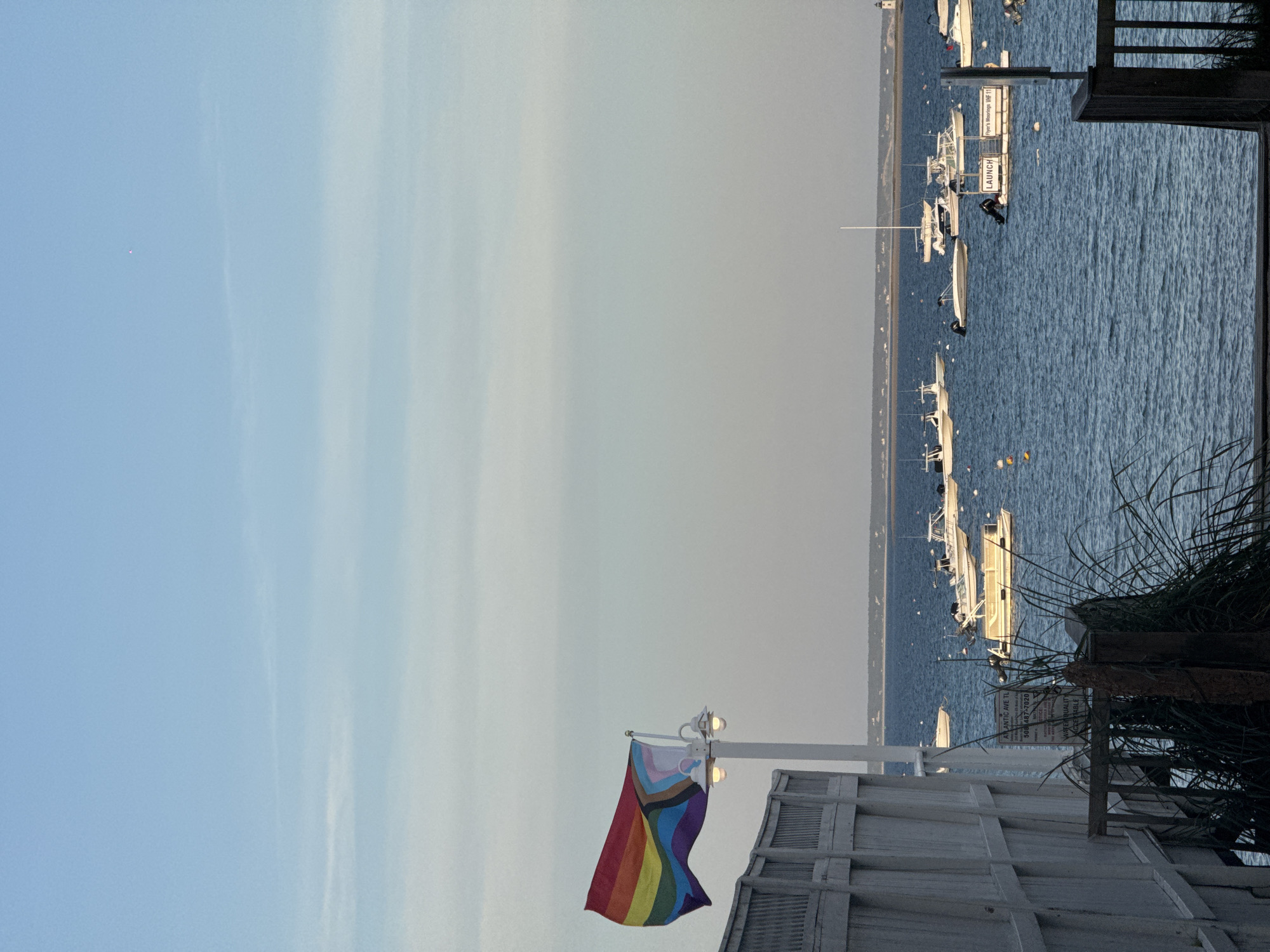 The image captures a tranquil coastal scene, likely a harbor or bay, under a bright, clear sky. In the foreground, a white wooden fence or railing partially frames the lower left, while dark green foliage fills the right edge. Prominently featured in the mid-ground is a flagpole with two streetlights attached, from which a vibrant Progress Pride Flag unfurls, displaying its traditional rainbow stripes along with a chevron of pink, light blue, white, brown, and black.
Beyond the foreground structures, a calm body of deep blue water stretches across the frame, populated by numerous boats of varying sizes, predominantly white. Many of the boats appear to be moored or docked, suggesting a busy maritime area. In the water, a floating dock or platform is visible, bearing the white text "LAUNCH" and "Hyer's Moorings WIF 11". A distant, low-lying landmass defines the horizon across the water. Amidst the foliage on the right, a partially visible sign contains text including "508 487-7030", "ATLANTIC AVE TL", and "NOTICE".
The sky transitions from a pale blue at the top left to a slightly hazy, lighter hue towards the right, with faint wisps of clouds present. A very small, bright pink dot is discernible high in the sky in the upper left. The lighting suggests either late afternoon or early morning, casting a soft, even glow over the scene. No people are visible, contributing to the serene atmosphere. The presence of the Progress Pride Flag and the maritime setting are consistent with the provided location context of Provincetown.