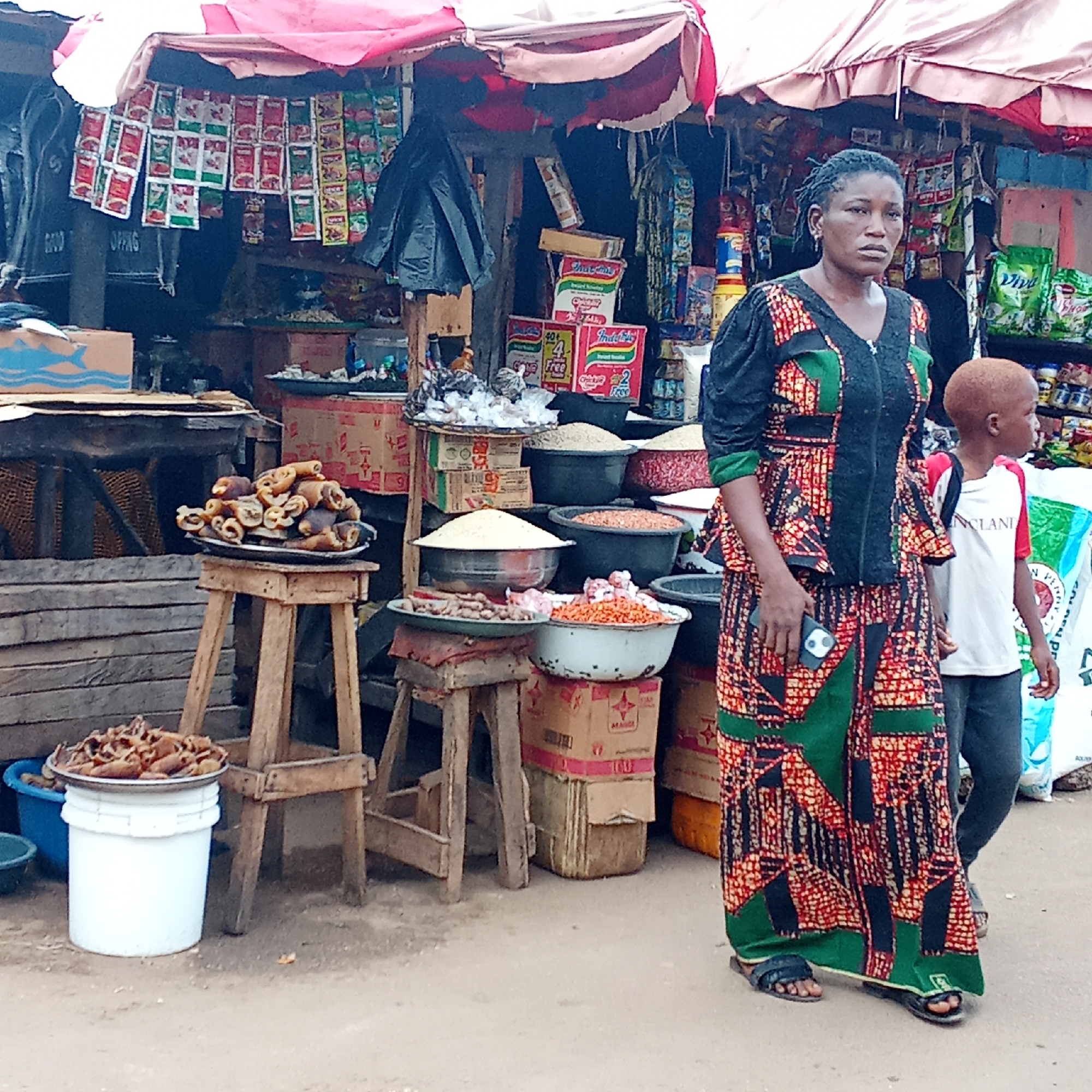In Akwanga, Nigeria, a woman in a colorful patterned dress and sandals stands next to a young boy with light hair, presumably her son. The woman is holding a smartphone. They are near a market stall under a striped awning, filled with food items such as grains in bowls, dried foods on trays atop wooden stools and a white bucket, and packaged goods. Boxes, sacks, and various containers support the arrangement of goods. The scene suggests a bustling market environment in Akwanga.