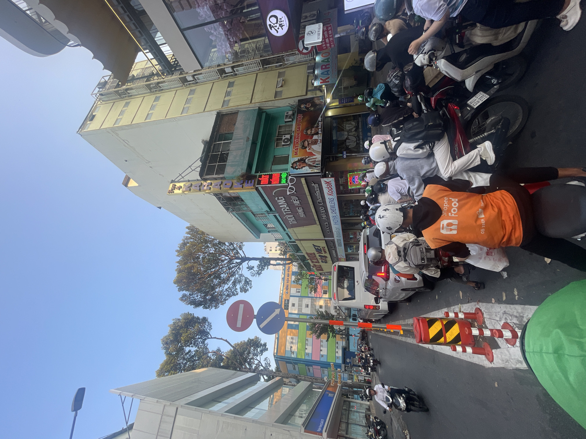 This image captures a vibrant and busy daytime street scene in Thủ Đức, Vietnam, characterized by a high volume of motorbike traffic under a clear, bright blue sky. The atmosphere is bustling, reflecting daily urban life.
The central subjects are numerous people on motorbikes, many wearing helmets, navigating what appears to be a busy intersection or congested road. A prominent figure in the foreground wears an orange uniform with "Shopee Food" and "CÓ TRÊN" visible, indicating a food delivery driver. Other vehicles, including a white van or mini-bus, are also part of the traffic flow.
The street is flanked by multi-story buildings housing various businesses. Storefronts display numerous signs in Vietnamese and English. Visible text includes "MẮT KÍNH SAO VIỆT" (Sao Viet Eyeglasses) advertising brands like "ESSILOR HOYA CHEMILENS Kodak" and offering "Chuyên gia kính độ" (Eyeglass specialist). Other signs indicate "THỜI TRANG" (Fashion), "HÀNG HIỆU" (Branded Goods), "KARAO" (Karaoke), and "SUSHI WAGAO." In the background, a colorful building is identifiable near a sign for "BỆNH VIỆN NHI ĐỒNG" (Children's Hospital).
Notable details include traffic signs such as a "no entry" sign and a "turn right/ahead" sign, along with red and white striped road barriers. Large green trees are scattered throughout the urban environment, breaking up the lines of buildings. The scene depicts a typical busy day in a Vietnamese city, with constant movement and commercial activity.