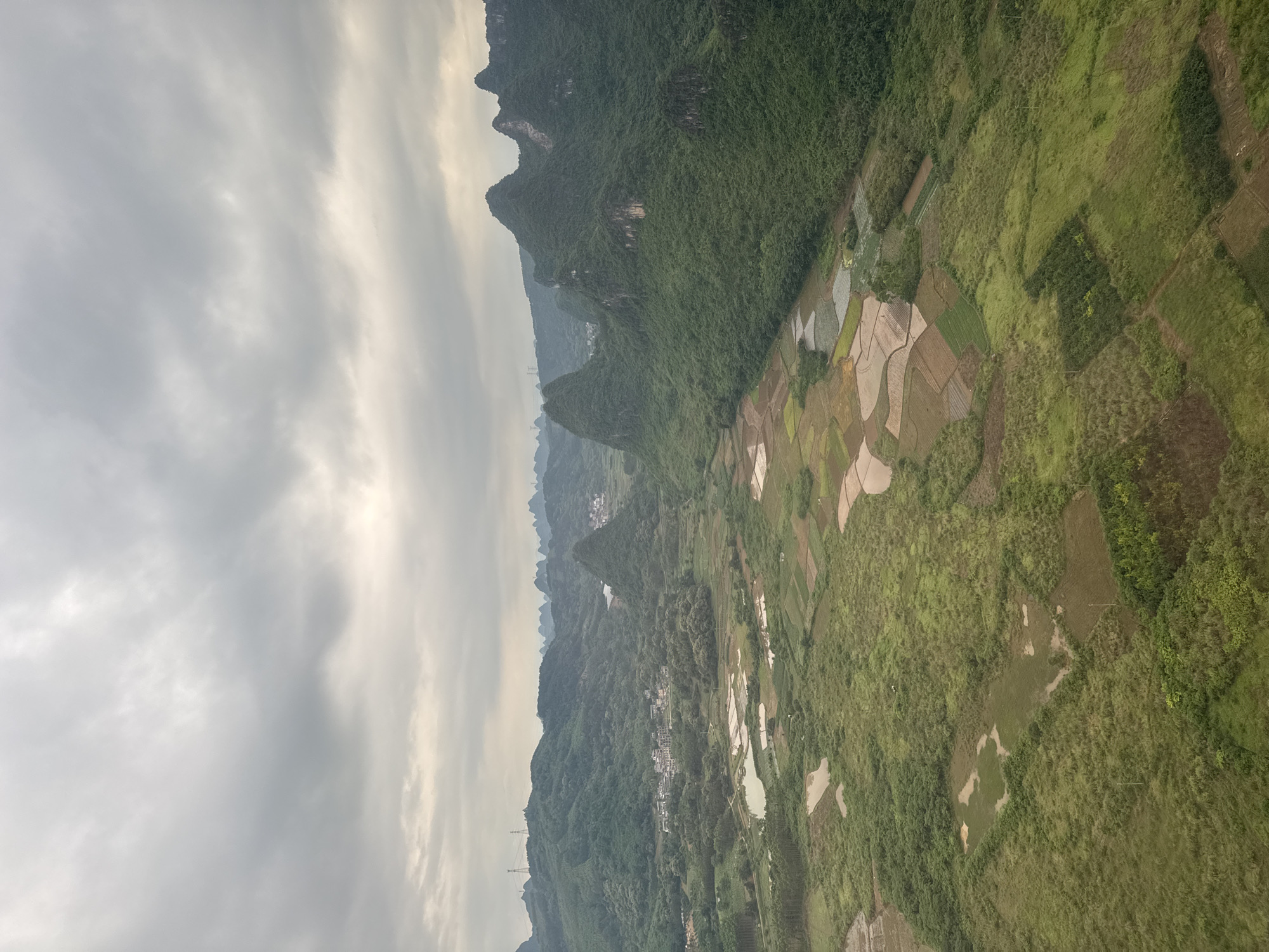 The image shows a landscape in Guilin, China. The scene features karst mountains densely covered with green vegetation. Rice paddies are visible in the valley, arranged in an organized pattern, some flooded with water, indicating active cultivation. The sky is overcast, with grey clouds obscuring the sun. The overall impression is one of a serene agricultural landscape set against a backdrop of striking natural formations.