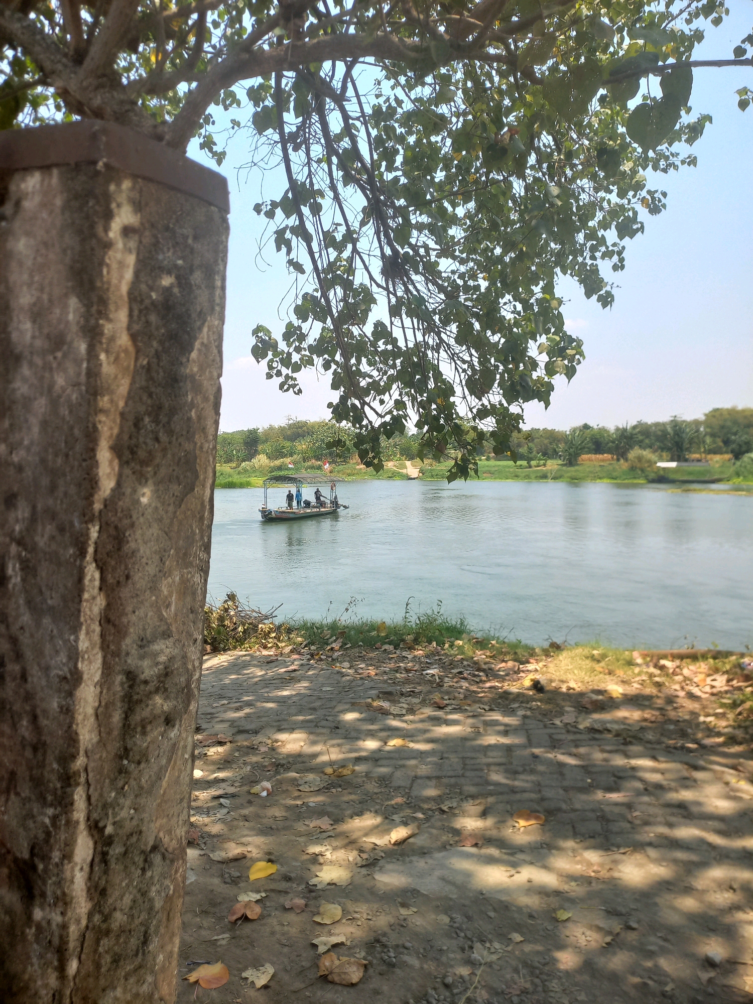 A body of water, likely a river, fills the right two-thirds of the frame. On the left, a rough-hewn stone pillar, stained and weathered, occupies the left third of the image. Branches of a tree with green leaves hang from the top of the frame, casting dappled shadows on a dirt and paved path in the foreground. On the water, a small boat with a canopy and several people on board is visible. The background features a treeline and a hazy sky. The location, Puskesmas Keboan, Indonesia, suggests this is likely a scenic view near the health center, possibly a recreational area.