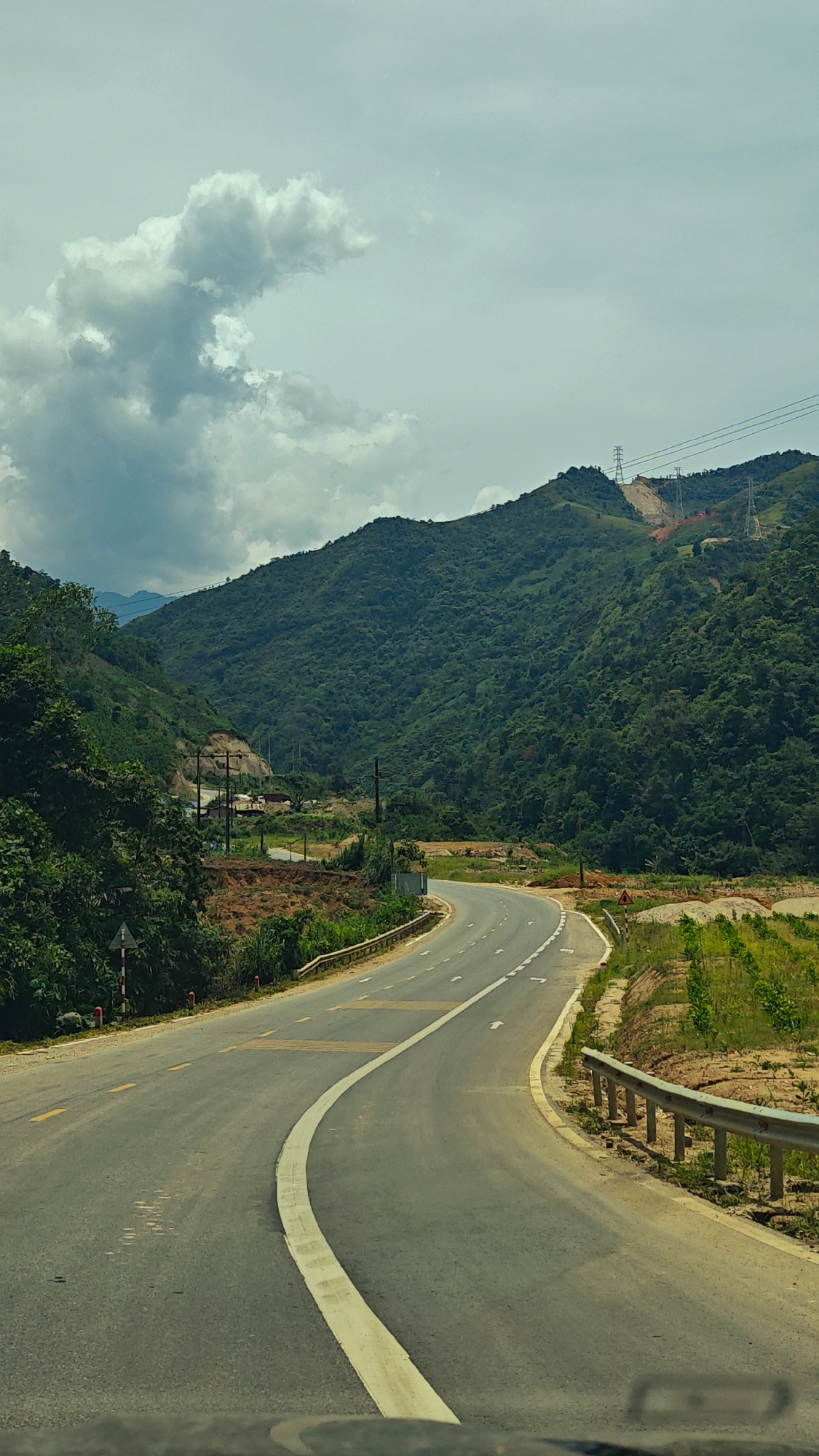 The image captures a scenic view of a winding road cutting through a lush mountainous landscape in Duong Quy, Vietnam. The asphalt road dominates the foreground, curving first to the right and then gently to the left as it recedes into the distance. It features a solid white line delineating the edge and a broken white line with directional arrows, indicating a path of travel. Further along, yellow dashed lines are visible on the road surface, possibly rumble strips or markings for a specific zone. A metal guardrail parallels the right side of the road in the foreground.
On both sides, dense, verdant mountains rise steeply, covered in abundant green foliage, suggesting a tropical or subtropical climate. The sky is overcast with a blanket of grey and white cumulus clouds, indicating a cloudy daytime, likely late morning or early afternoon. In the mid-ground, a valley opens up on the left side, revealing some sparse human settlements and utility poles nestled among the trees. On the right side of the road, beyond the guardrail, there's a cultivated field with young green plants arranged in neat rows.
Further into the background, several tall power pylons carrying electrical lines stretch across the mountain ridges. Portions of the mountain slopes appear to have been cleared, showing exposed reddish-brown earth, possibly for infrastructure development related to the power lines. Two road signs are visible: a small red-and-white sign on the left edge of the road and a yellow triangular warning sign on the right. No people or other vehicles are explicitly visible, suggesting a tranquil and possibly remote setting, with the perspective taken from inside a moving vehicle.