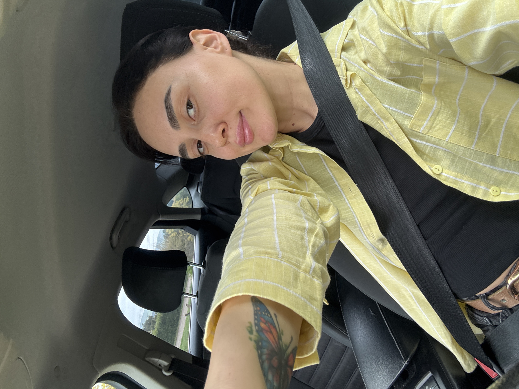 A close-up, tilted selfie captures a young person seated in the passenger seat of a car, looking towards the camera with a soft smile. The subject has dark hair pulled back, prominent dark eyebrows, and light makeup, giving them a natural appearance. They are wearing a light yellow long-sleeved button-up shirt with thin white vertical stripes over a black top, and a brown belt is partially visible. A black seatbelt crosses their chest and lap.
A colorful butterfly tattoo, featuring shades of blue, orange, and green, is visible on their right forearm, which is extended towards the camera. The car interior features dark seats with visible stitching and dark headrests. Through the car windows, a bright, possibly overcast, daytime sky and green foliage with distant landscape features, including what appears to be a road and possibly power lines, can be seen, indicating the vehicle is in motion or parked outdoors. The overall setting is a car in Tbilisi, Georgia. No visible text is present in the image.