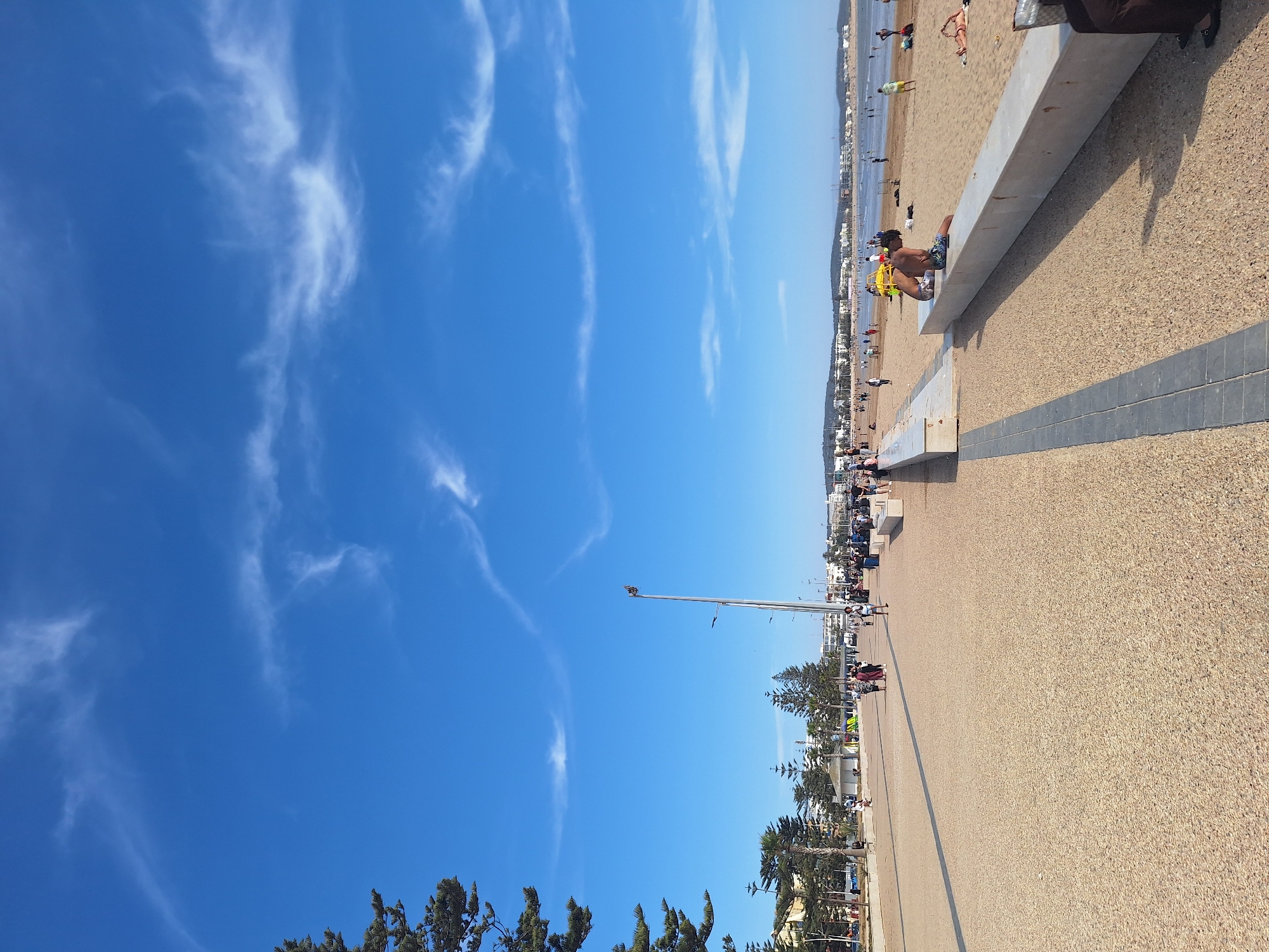 The image depicts a sunny day at the beach promenade in Essaouira, Morocco. The promenade is paved with light brown aggregate and features a dark grey border. To the right of the promenade is a sandy beach where several people are visible, some sunbathing and others walking. A long, low concrete bench runs along the edge of the promenade, with a few individuals seated on it. In the distance, the ocean is visible, with waves gently rolling onto the shore. Further back, the cityscape of Essaouira can be seen, with buildings and trees dotting the landscape. The sky is a clear, bright blue with a few wispy white clouds. Tall pine trees are present on the left side of the image, framing the view. A lamppost stands tall along the promenade, and a flag is attached to it. The overall scene conveys a relaxed and pleasant atmosphere, typical of a seaside resort.