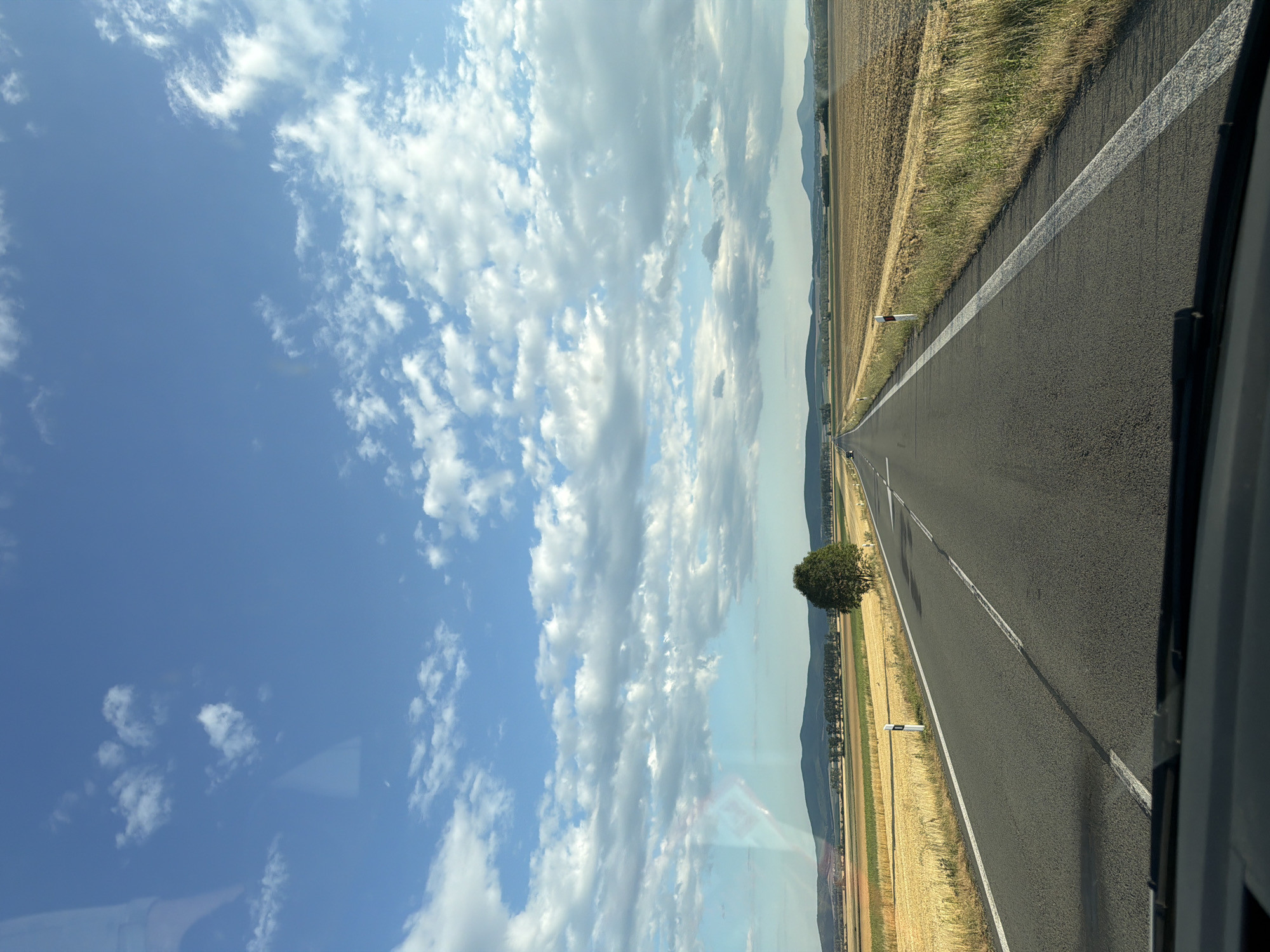This is a horizontal, eye-level, medium shot capturing a road leading into the distance through a rural landscape in Zemplínske Hradište, Slovakia. The road is asphalt with white lane markings and reflective posts on the right side. On either side of the road are fields, one appearing to be harvested grain, and the other dry, brown grass. In the middle ground, a single, mature tree stands to the right of the road. The background features rolling hills and mountains under a bright blue sky filled with scattered, fluffy white clouds. The foreground shows the dashboard and windshield wipers of a vehicle, indicating the photo was taken from inside a car. The scene is bathed in natural daylight, suggesting it was taken during the daytime. There are no people or animals visible in the frame.