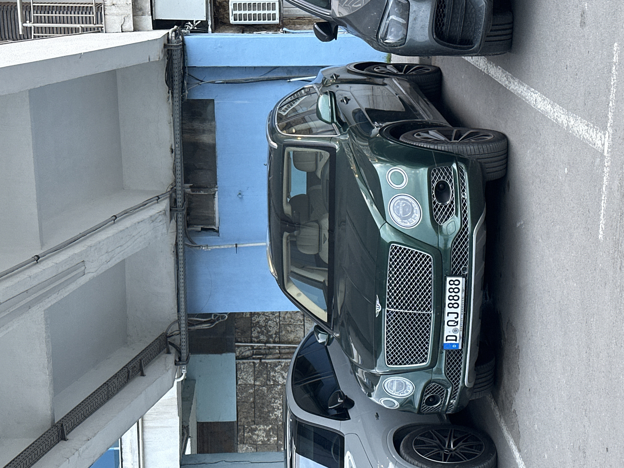 The image, rotated to a vertical orientation, features a dark green Bentley Bentayga SUV parked on an asphalt surface. The vehicle is centrally placed, displaying its large, mesh grille, distinctive circular headlights, and multi-spoke alloy wheels. A German license plate, "D QJ 8888," is prominently displayed on the front, with the text "BENTLEY HANNOVER" visible beneath it, indicating its origin or dealership.
To the right of the Bentley, the front portion of another dark grey SUV is partially visible, while to the bottom-left, the front wheel and bumper of a lighter grey car can be seen, suggesting a row of parked vehicles. Faded white lines mark the parking spaces on the asphalt.
The background consists of architectural elements. A light-colored building structure with concrete beams and a textured white wall frames the left side of the image. Numerous conduits and cables are affixed to these structures. Directly behind the Bentley is a wall painted a bright blue, providing a contrasting backdrop. Above this blue wall, a small, louvered white ventilation unit is visible.
The scene takes place during the daytime, under what appears to be an overcast sky, resulting in soft and even illumination. No individuals are present in the frame. The visible German license plate and "Bentley Hannover" text suggest a location in Germany, which contradicts the provided context of Varna, Bulgaria.