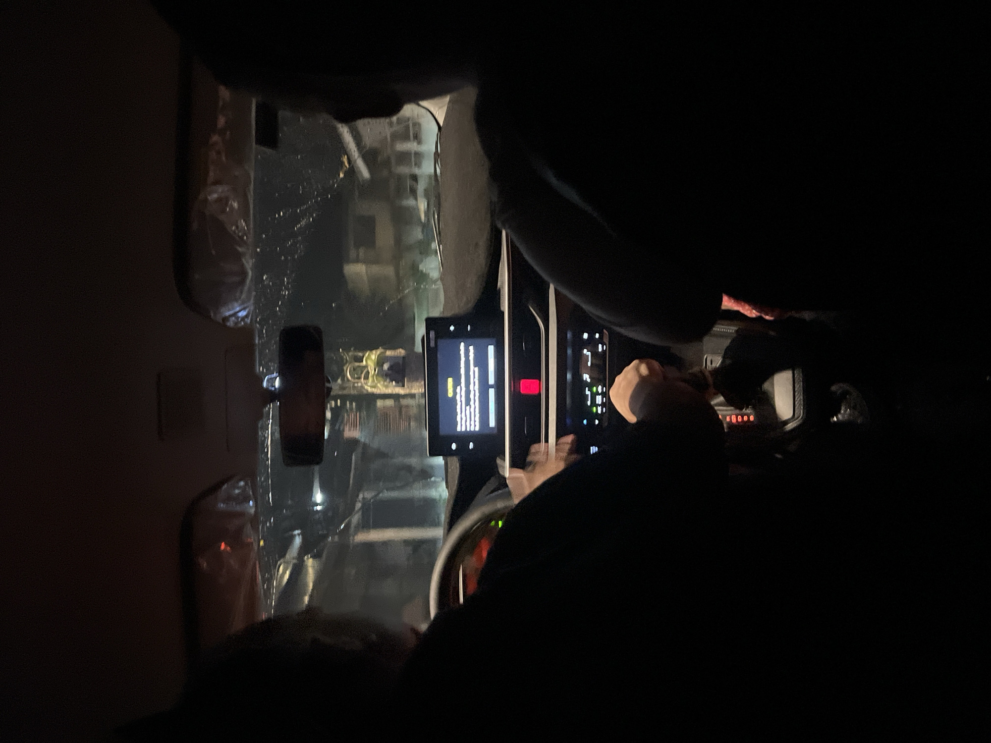 The image shows an individual in a car at night in Davao City, Philippines. The interior of the car is visible, with a lit-up dashboard and central display. The windshield shows a blurred view of city lights and buildings outside. The individual's hand is on the steering wheel. A mobile phone is visible on the dashboard.