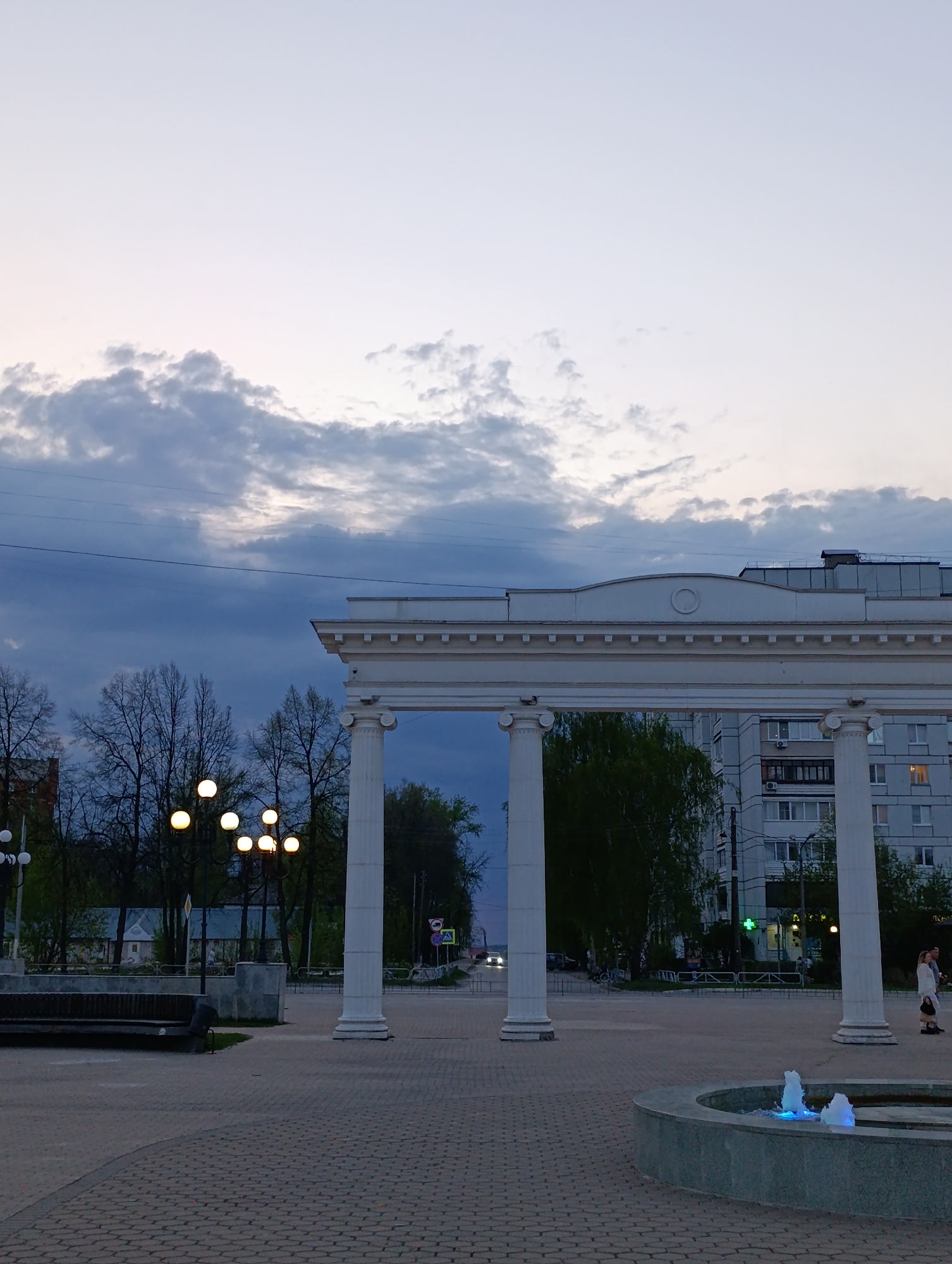 The image depicts a scene in Chaikovsky, Russia. In the foreground is a paved square with a small fountain illuminated by blue lights. Behind the fountain stands a series of white pillars connected by a white arch, forming a colonnade-like structure. Through the archway, a street with traffic is visible. To the left, there is a park area with bare trees and illuminated spherical lamps. In the background, a multi-story building is present. The sky above is a mix of gray and pale blue with scattered clouds, suggesting twilight or dusk. A few people are walking on the paved area.