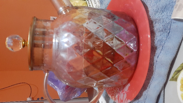 An indoor, close-up image focuses on a clear glass teapot with a faceted diamond pattern on its body, C-shaped handle, and spout. The teapot contains a reddish-brown liquid. Its clear lid features a gold-colored band and a faceted clear glass knob. The teapot rests centered on a solid red-orange ceramic plate adorned with a partial white floral design along its rim. This plate is positioned on a light blue terry cloth towel. The background consists of a plain orange wall, with a white electrical outlet and black wires visible on the upper left, and a blurred purple plastic bag behind the teapot's handle. To the right, another blurred, light-colored object and patterned fabric are partially visible. In the immediate foreground, a portion of a white plate with painted yellow lemon slices and green leaves is seen, alongside a sliver of white packaging. No people are present. Specific identifying features for the city of Pap, Uzbekistan, are not discernible within the image.