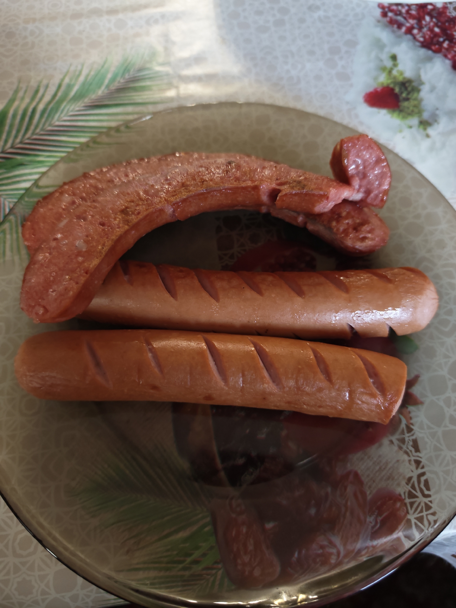 Three sausages are placed on a decorative plate with a palm leaf pattern. The top sausage is split and curved, with one piece overlapping another. It appears to be cooked and slightly browned, with some visible spices. The other two sausages are whole and appear to be uncooked, with diagonal scores cut into their surfaces. They are positioned one below the other, with the top one slightly to the left of the bottom one. The plate is on a tablecloth with a light-colored patterned design that includes what looks like small red berries. The lighting suggests it might be daytime, possibly indoors. The overall impression is a close-up shot of food, likely prepared or about to be prepared.