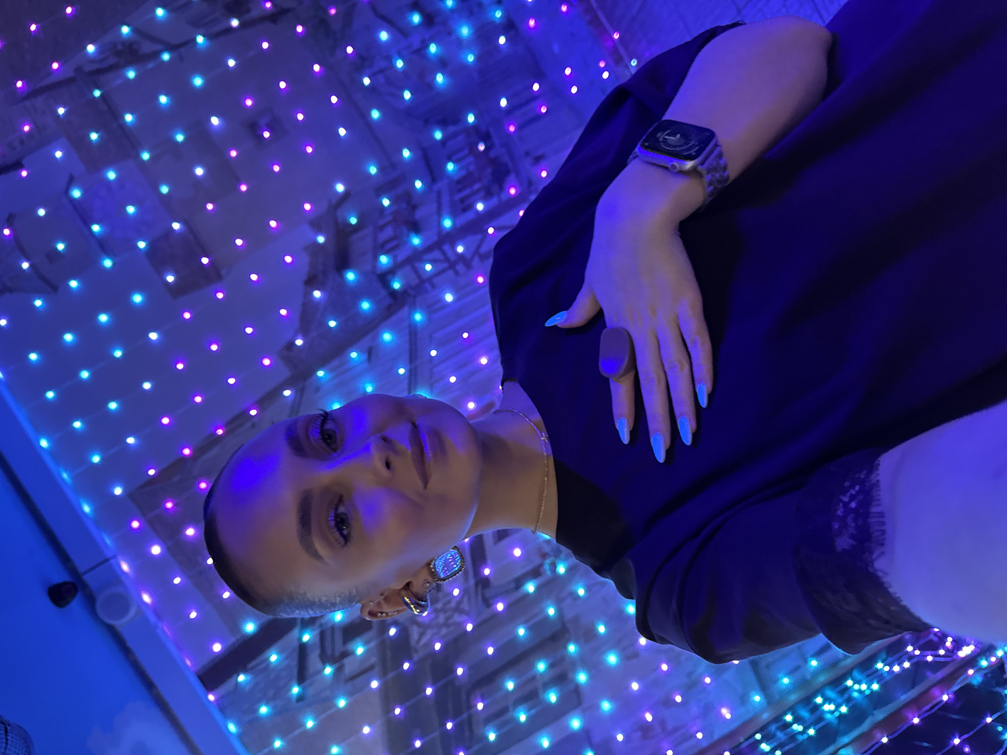 A woman is captured in a selfie within an indoor establishment, likely an entertainment venue in Chaikovsky, Russia. The background features a wall illuminated by numerous small LED lights arranged in a grid pattern, displaying alternating cyan and magenta hues. Partially visible behind these lights is a mural depicting stylized architectural forms, possibly buildings or castles.
The subject is a woman with light skin and dark hair pulled back tightly. She wears subtle makeup, including winged eyeliner, and exhibits a faint smile directed towards the camera. Her left ear is adorned with multiple pieces of jewelry: a small helix piercing, a curved metallic earring, and a prominent rectangular dangle earring featuring a light-blue or iridescent stone. A delicate gold chain necklace is visible around her neck. She is dressed in a dark, possibly black, short-sleeved top with a visible lace trim on the cuff of her right sleeve. Her fingernails are painted a bright sky-blue. Her left hand, wearing a large, dark, possibly wooden ring on the ring finger, rests gently on her chest. A smartwatch with a patterned band is worn on her right wrist. Overhead, in the upper left portion of the image, a small black surveillance camera and a white circular fixture (possibly a speaker or smoke detector) are mounted on the ceiling. The entire scene is bathed in the predominant blue and purple light emitted by the LED display. The precise name or address of the establishment within Chaikovsky cannot be determined from the image.