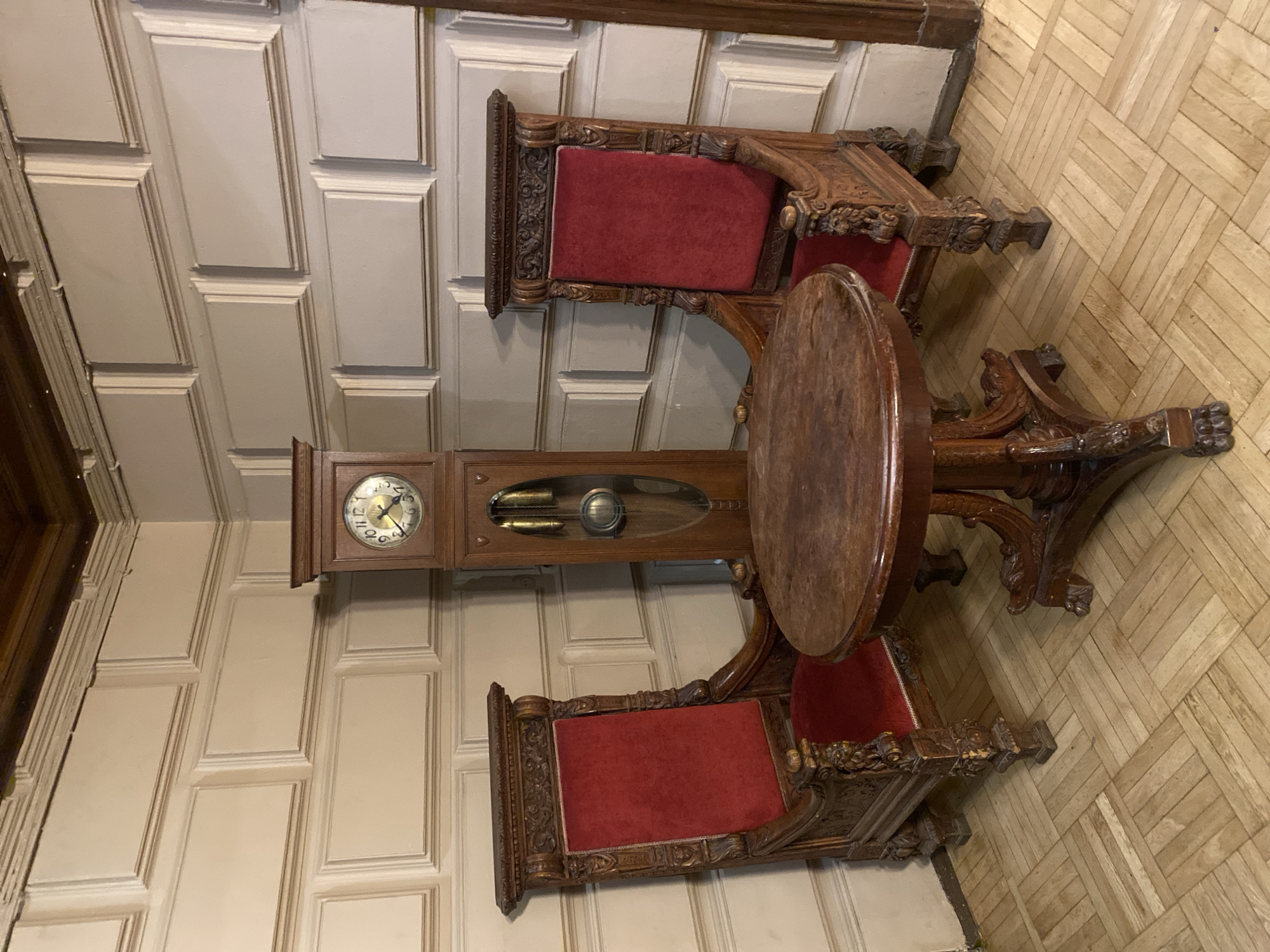 The image depicts an interior scene with a grandfather clock positioned between two ornate chairs. A round wooden table stands nearby. The chairs are upholstered with red fabric. The room has paneled walls and a parquet floor. The scene does not provide enough information to determine a specific location in Moscow, Russia, or anything about the people or actions happening in the room.