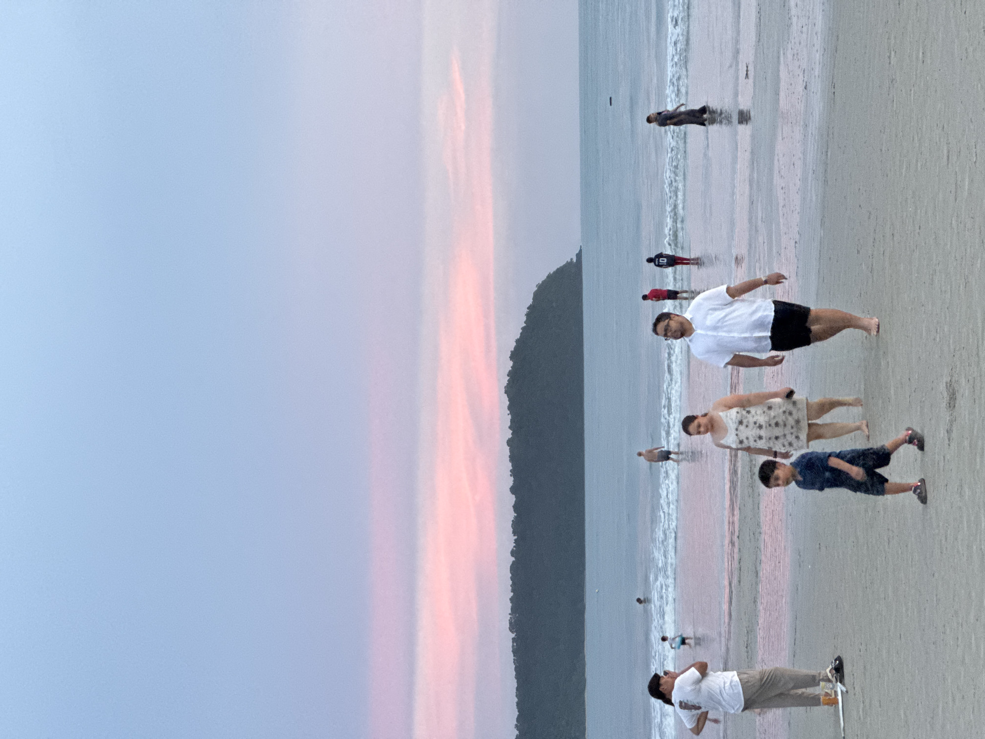 The image depicts a beach scene at dusk on Cenang Beach, Malaysia. The sky transitions from a pale blue at the top to soft pink and lavender hues near the horizon, indicating sunset. A dark, tree-covered hill or island is visible in the distance, separating the sea from the sky. The ocean is calm with gentle waves breaking on the shore. The sandy beach is wet and reflective near the water's edge.

Several people are present in the scene. In the foreground, a young boy in a dark blue t-shirt and shorts walks towards the right. To his left and slightly behind, a woman in a floral print dress walks with her head turned. Further to the left, a man in a white t-shirt and light-colored pants stands with his back to the camera, looking towards the sea. In the mid-ground, a man in a white button-down shirt and black shorts walks towards the left, with his bare feet in the shallow water. To his right and further back, a person in a red t-shirt and another in a dark shirt with the number '10' are visible in the water. On the far right, a person in a dark dress stands in the water. In the background, a few other figures are scattered in the sea and on the beach. The overall atmosphere is serene and relaxed, typical of an evening at the beach.