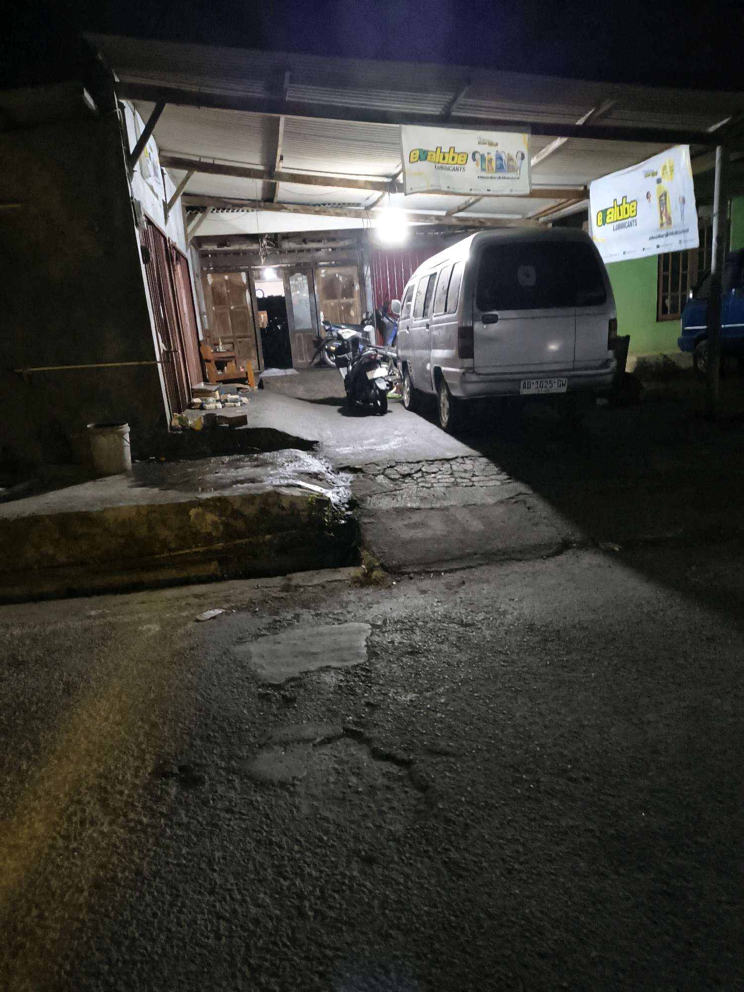 The image depicts an outdoor scene at night, likely in Randugunting, Indonesia. In the foreground, a wet, asphalt road shows signs of disrepair with puddles and cracks. A concrete curb runs along the left side of the road, with a metal bucket placed on it.
Further back, a light-colored van is parked. Its license plate reads "AD-1025-GW". To the left of the van, a motorcycle is parked. Above and behind the van, two banners are hung, featuring the "evalube" logo and images of lubricant products. The banners suggest a commercial or promotional setting.
The background reveals a building with an open-fronted structure, possibly a shop or workshop, with wooden doors and interior lights. Another blue vehicle is partially visible on the right side of the image. The overall environment appears somewhat unkempt, with evidence of wear and tear on the infrastructure. The lighting is dim, with artificial lights from the building and possibly from streetlights creating shadows. The atmosphere is that of a typical street scene in a residential or commercial area at night.