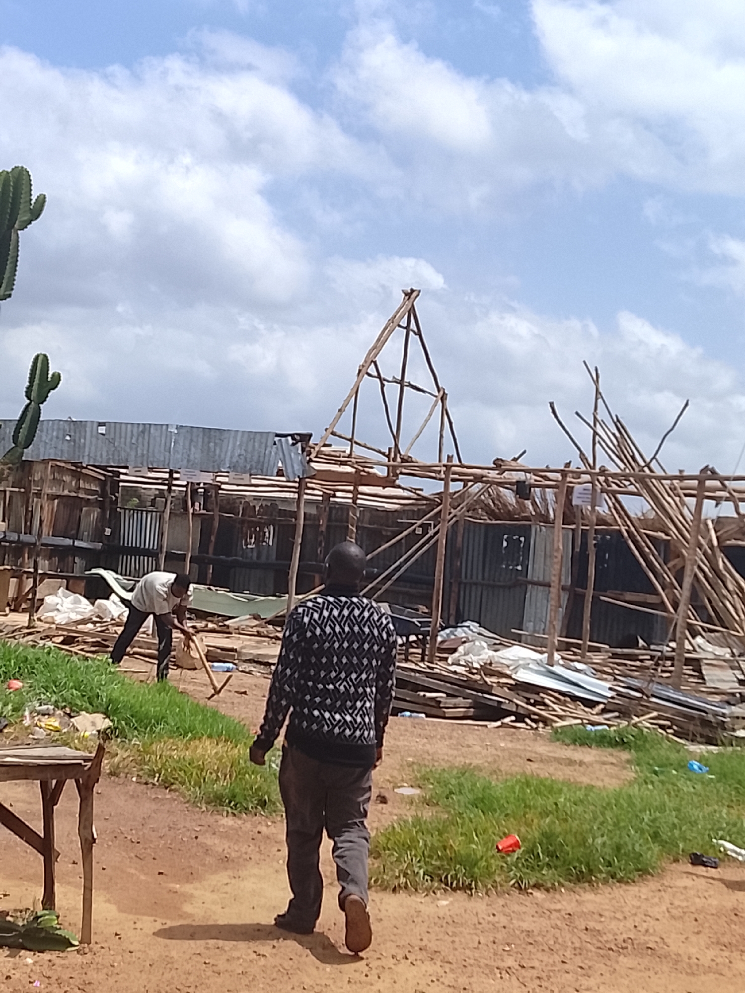 The image shows an outdoor scene with two men and a partially constructed or damaged wooden structure. In the foreground, a man is walking away from the camera, wearing a black and white patterned shirt and grey pants. To the left, another man is bent over, working with a tool that appears to be a shovel or hoe. He is wearing a light-colored shirt and dark pants.
The background features a structure made of wood and corrugated metal, appearing in a state of disrepair or unfinished construction. Scattered materials, including wood planks and white sheeting, are visible around the structure. A large cactus is prominently displayed on the left side of the image, with a smaller one below it.
The ground is a mix of dirt and patchy green grass. There is some litter on the grass, including a red cup and a blue plastic bag. The sky is cloudy, with patches of blue sky visible, suggesting it might be daytime with varied weather. The overall impression is one of manual labor, possibly construction or demolition, in an unrefined outdoor setting. No discernible text is visible in the image.