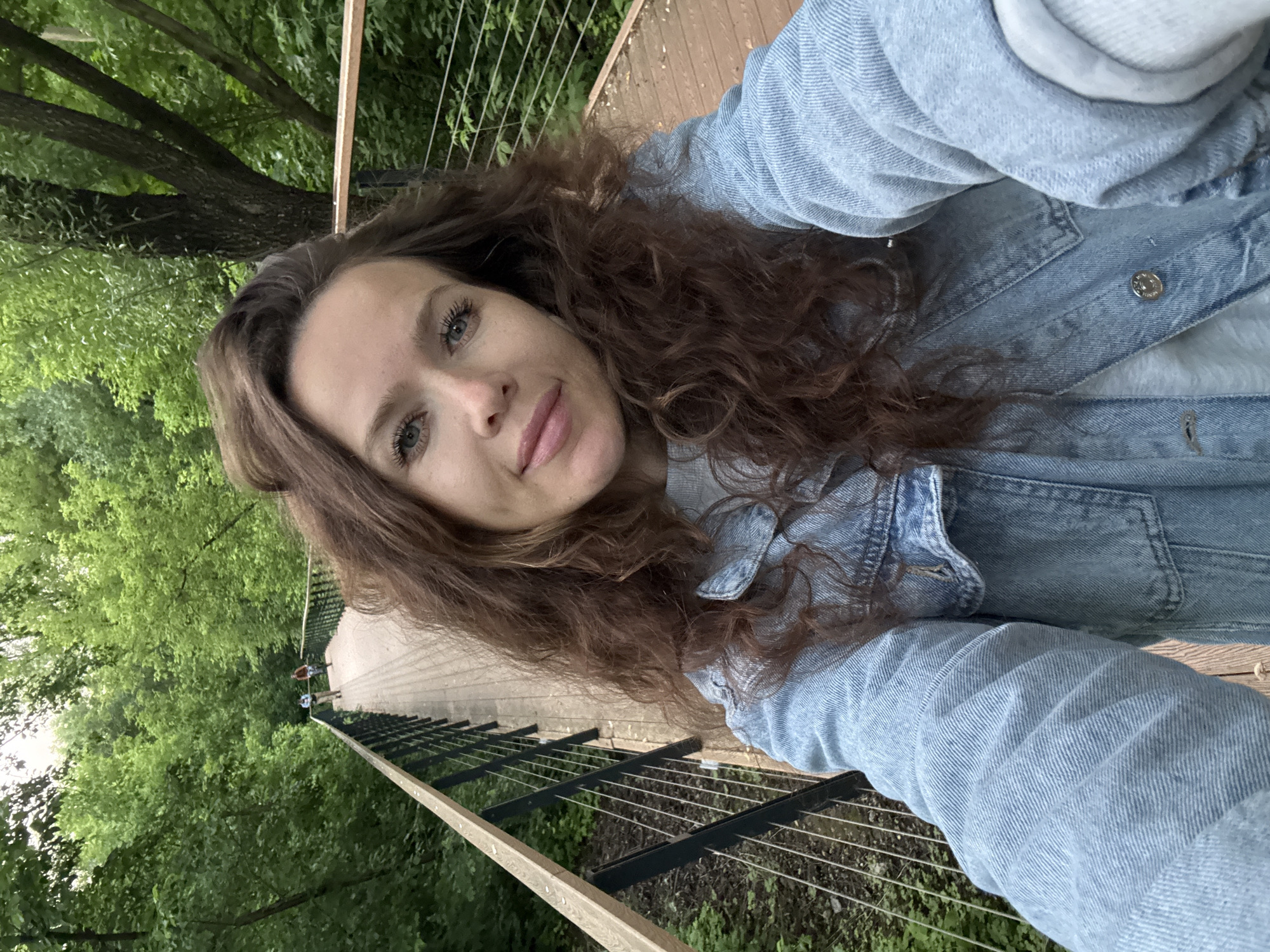 A woman with long, curly brown hair and blue eyes is taking a selfie on a wooden walkway in a park. She is wearing a light blue denim jacket over a gray sweater. The walkway has a dark metal railing with thin wires. In the background, two people can be seen walking away from the camera on the same walkway, which is surrounded by lush green trees. The scene appears to be outdoors in a natural setting.