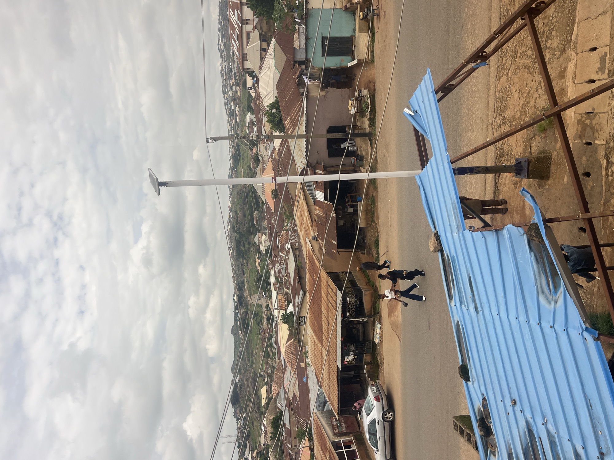 The image shows an aerial view of a street in the city of Jos, Nigeria. The street is lined with buildings with corrugated iron roofs, some of which appear to be in disrepair. There are several people walking on the street and on the sidewalks. A silver car is parked on the side of the road. In the foreground, a partially constructed structure with a blue corrugated iron roof is visible. A solar-powered street light stands on the sidewalk. The background features a hilly landscape with more buildings scattered across it. The sky is overcast with clouds.