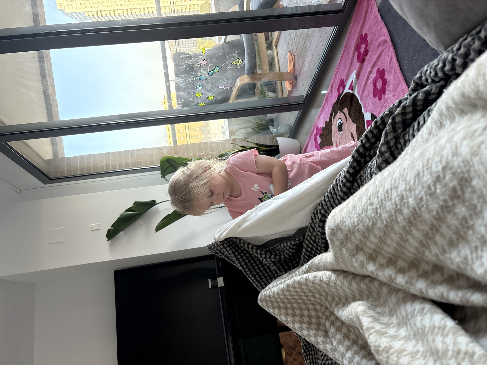 A young blonde girl, wearing a pink t-shirt and shorts, is standing in a living room in Valencia, Spain. She is looking down at a blanket fort that she is building. The fort is made of several blankets and pillows, and it is draped over a sofa. A large window overlooks a balcony with a chair and plants. The floor is covered with a pink mat with a cartoon character on it. The apartment appears to be high up, as buildings can be seen in the distance.
