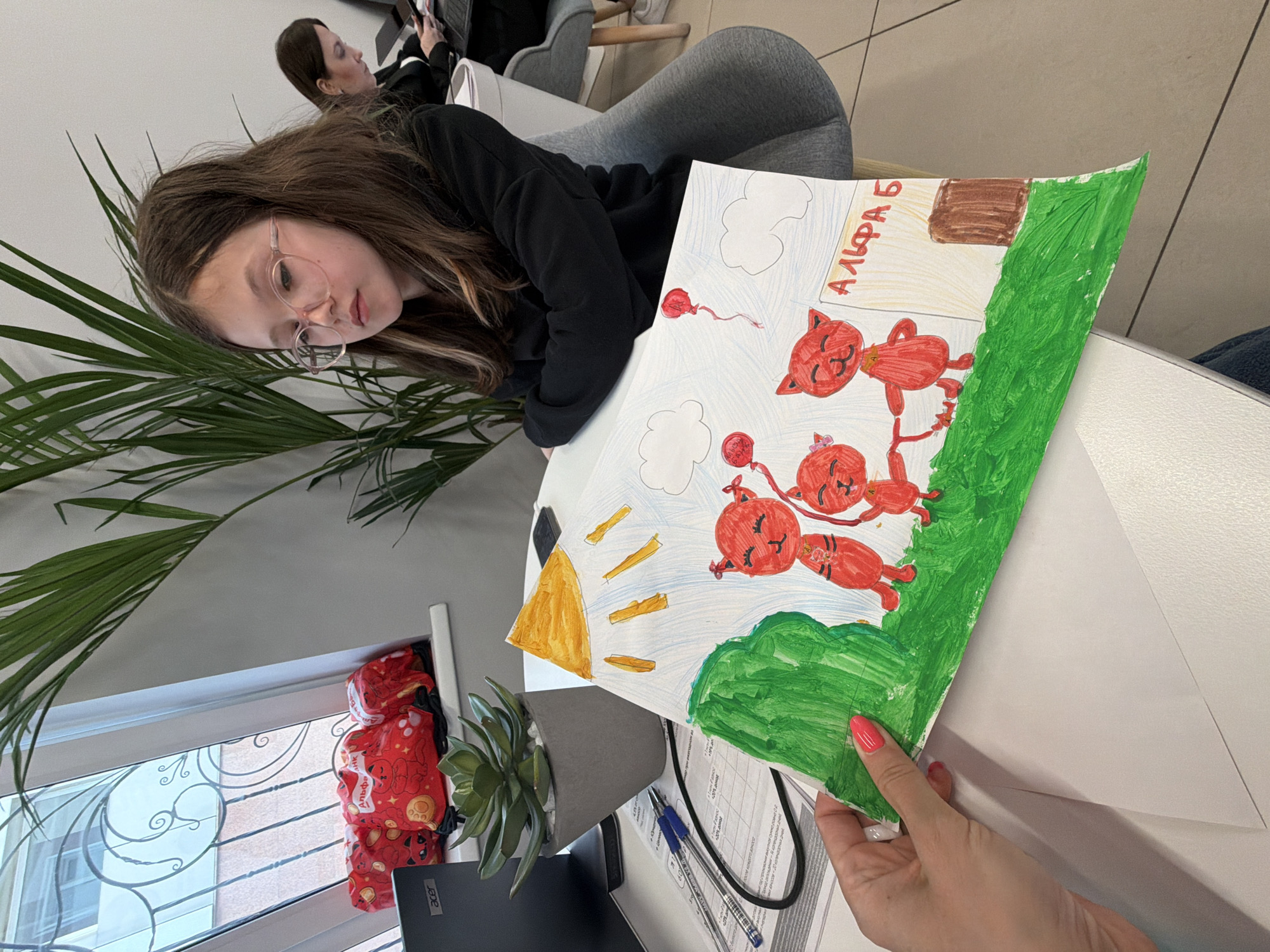 A medium shot captures a young girl with long brown hair and glasses, slightly tilted to the side, as she looks at a drawing. The drawing, held by an adult hand with manicured pink nails, depicts a colorful scene with red cats, green foliage, and what appears to be a house. The Russian word "Алфавит" (Alphabet) is written at the top of the drawing. The scene takes place indoors, as evidenced by the presence of a plant, a laptop, and other office supplies on the desk. Behind the girl is a window with a red bag hanging nearby and another person seated in the background. While the location is noted as Birobidzhan, Russia, there are no elements in the image that directly confirm this specific geographic location.