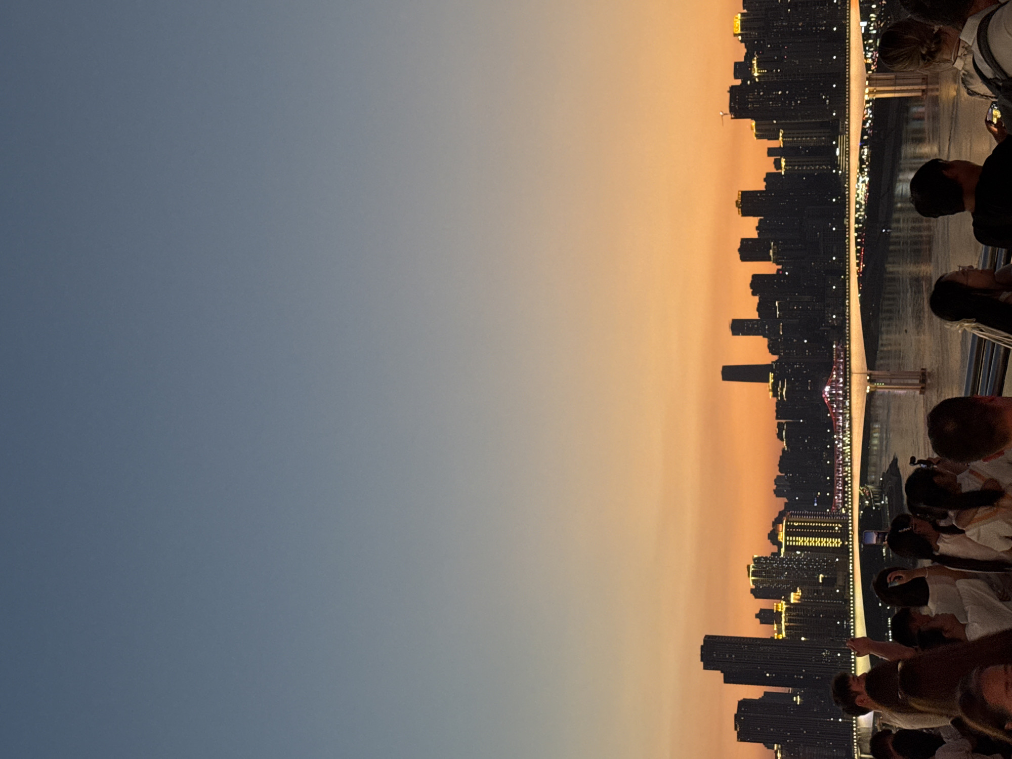 The image depicts a sunset scene in Yuzhong District, China, with a crowd of people on a walkway overlooking a cityscape and a river. The sky transitions from a pale blue at the top to a vibrant orange near the horizon. Silhouetted against the sunset are numerous tall buildings forming a city skyline, with some buildings illuminated by golden lights. A long bridge with red lighting stretches across the river, reflecting on its surface.
In the foreground, a diverse group of people are gathered, many looking towards the cityscape or holding up their phones to take pictures or record videos. The individuals are dressed in casual attire, suggesting a leisurely outing. Some are engaged in conversation, while others are observing the view. The atmosphere appears to be one of shared enjoyment and appreciation of the urban panorama at dusk. The walkway itself is lined with a railing, indicating it is a public observation area. The river is wide, and its surface is calm, reflecting the lights from the city and the bridge.