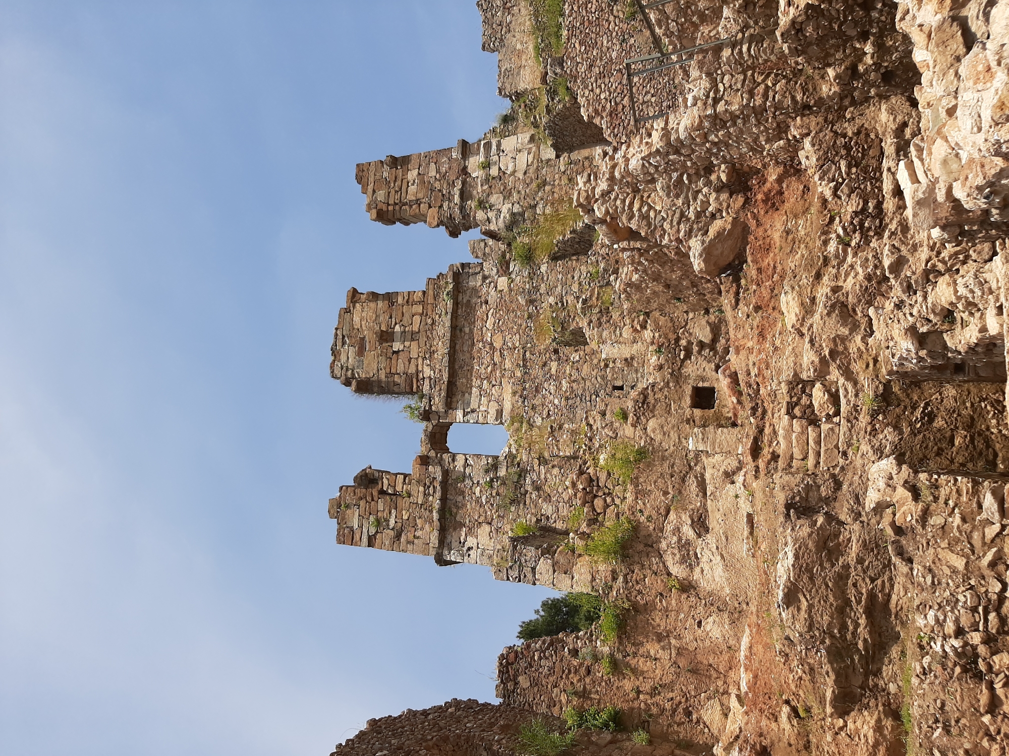The image captures a partial view of the ruins of Alanya Castle in Alanya, Turkey, set against a clear blue sky. The ruins consist of weathered stone walls and structures, showing various stages of decay with vegetation growing on and around the stones. The walls are built into a natural rock formation, which is visible alongside the stone structures. A metal railing is present along the edge of the ruins, suggesting a designated pathway for visitors. There are no people visible in the frame.