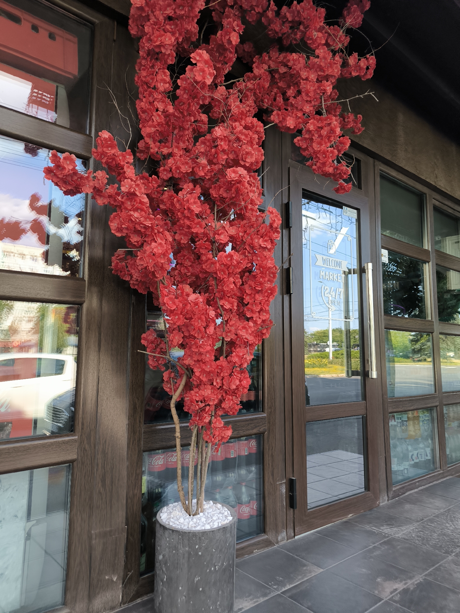 This is a medium shot of a storefront in Chisinau, Moldova. A large artificial red floral arrangement is prominently displayed in a gray planter pot filled with white stones. The arrangement is to the left of a double glass door with wooden framing. The door on the right has a handle and a sign that reads "WELCOME MARKET 24/7 OPEN." Reflected in the glass doors are the sky, buildings, and trees. Behind the glass, shelves stocked with Coca-Cola bottles are visible. The ground in front of the storefront is paved with dark gray square tiles.