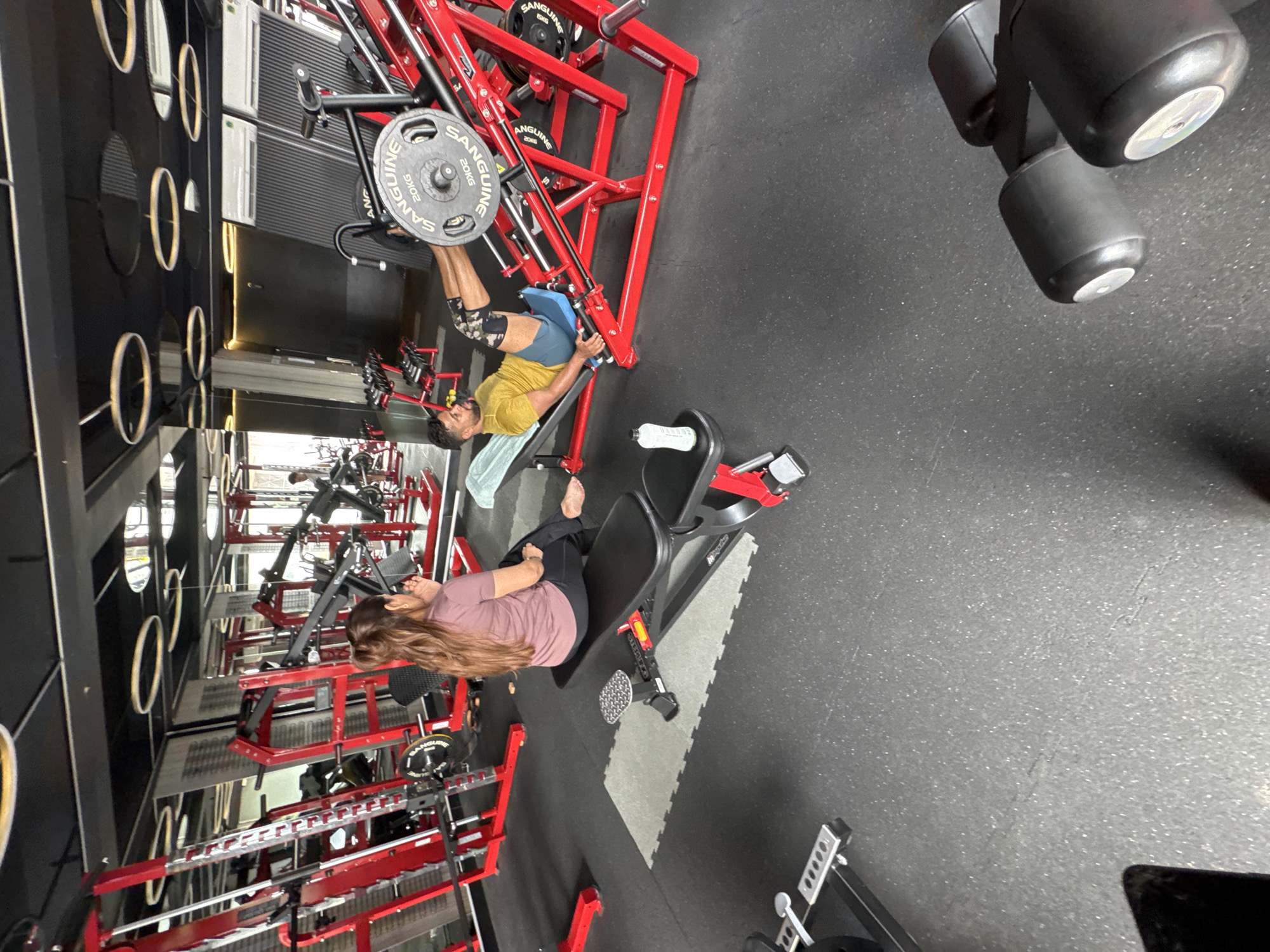 A gym scene is captured with two people, a man and a woman, engaged in different exercises. The man is using a leg press machine with "SANGUINE" branding. He is wearing a yellow shirt and blue shorts with knee support. The woman is using a seated leg curl machine and is wearing a mauve t-shirt and black pants. The gym appears to be well-equipped with various exercise machines. The walls are black, and there are circular decorations. There's a rack on the left side of the image. The location is not discernible from the image.