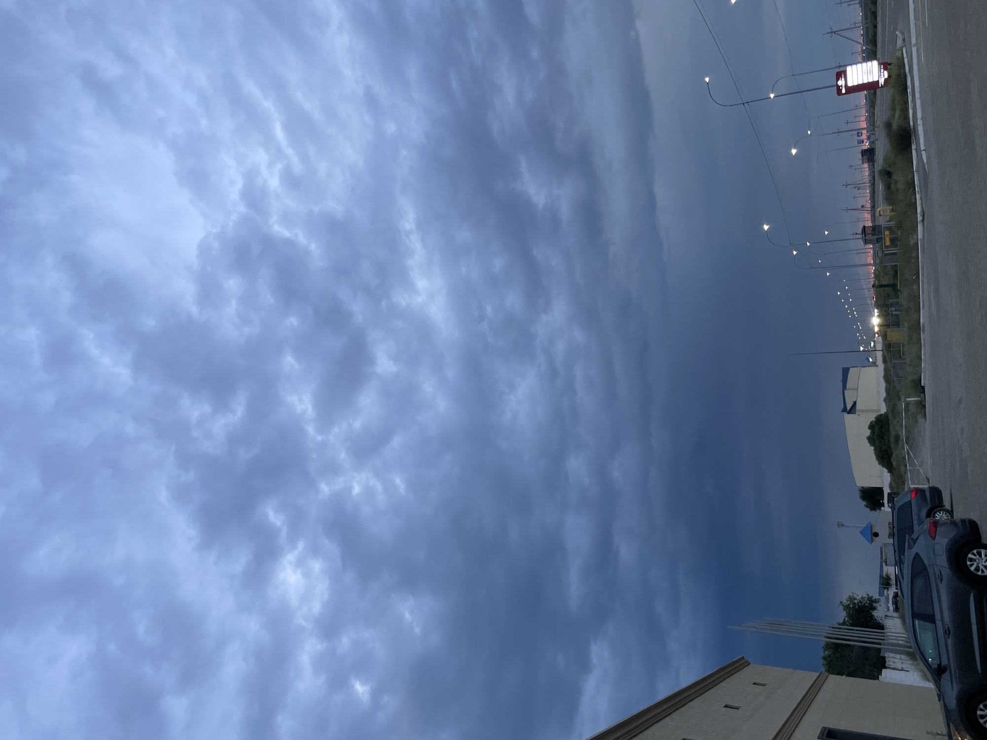 The image depicts a cloudy sky over an industrial area in Aktau, Kazakhstan. The clouds are dark and heavy, suggesting an impending storm or twilight. In the foreground, a grey car is parked in a paved lot. To the left, a beige building with rectangular windows is visible. Further in the background, a row of streetlights illuminates a road that curves into the distance, with more lights and buildings lining the route. The overall scene conveys a sense of an overcast day in a utilitarian, possibly commercial or industrial, part of the city.