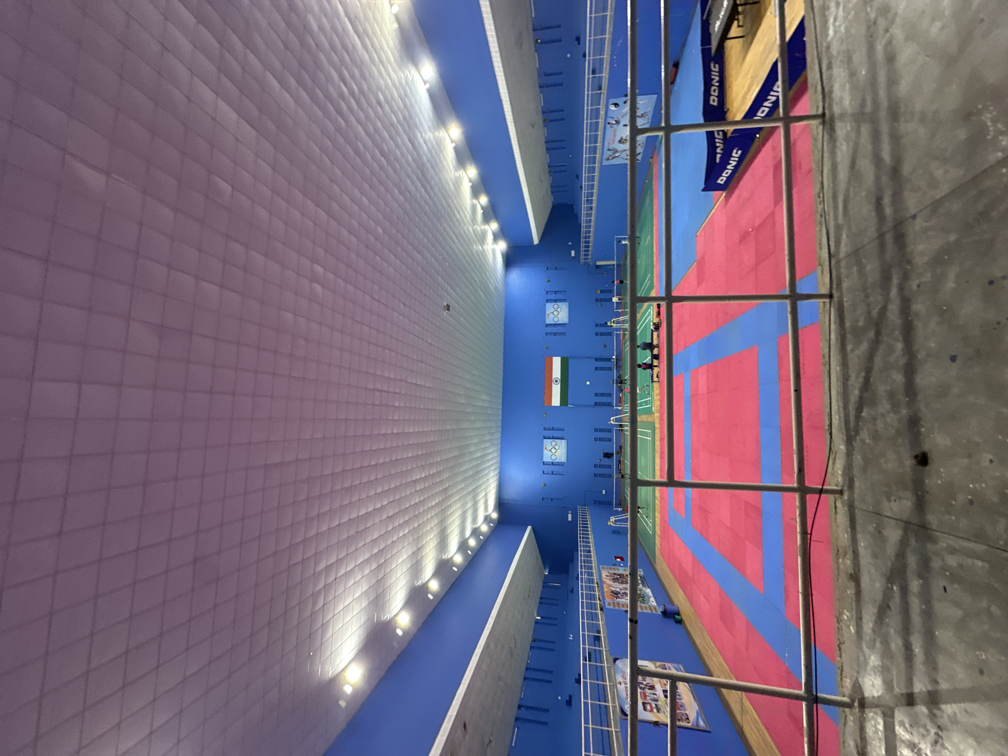 An indoor sports facility, likely a gymnasium, is depicted. The walls are painted a vibrant blue. The ceiling is a white, textured paneling with an integrated lighting system. The floor is marked for various sports, with sections of red and blue mats, and a green court area. In the background, there are badminton nets set up. An Indian flag is visible on the wall, along with Olympic rings. There are a few people present on the courts, some appearing to be playing or practicing. A railing in the foreground suggests the image was taken from a raised viewing area or walkway. The lighting is artificial and appears to be the primary illumination source.