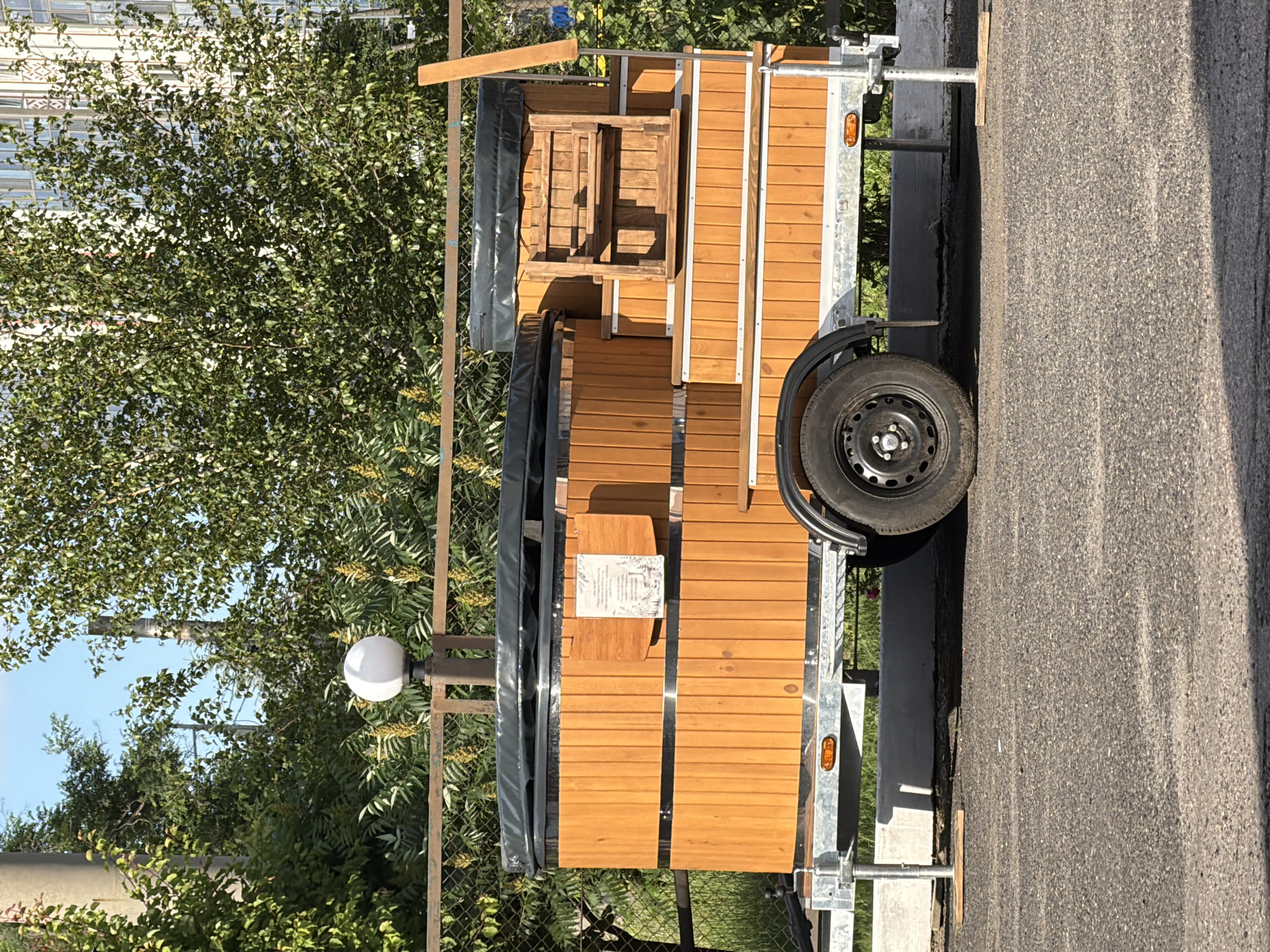 A wooden hot tub on a trailer is parked on an asphalt surface outdoors. The hot tub is circular and appears to be made of light-colored wood planks with metal bands. It has a black cover. A wooden shelving unit is attached to the side of the trailer, next to the hot tub. The trailer has a single wheel visible on the right side. In the background, there is a chain-link fence, trees with green foliage, and a white spherical lamp on a pole. The sky is clear and blue. The image is taken from a slightly low angle, looking up at the hot tub. There are no people visible in the image.