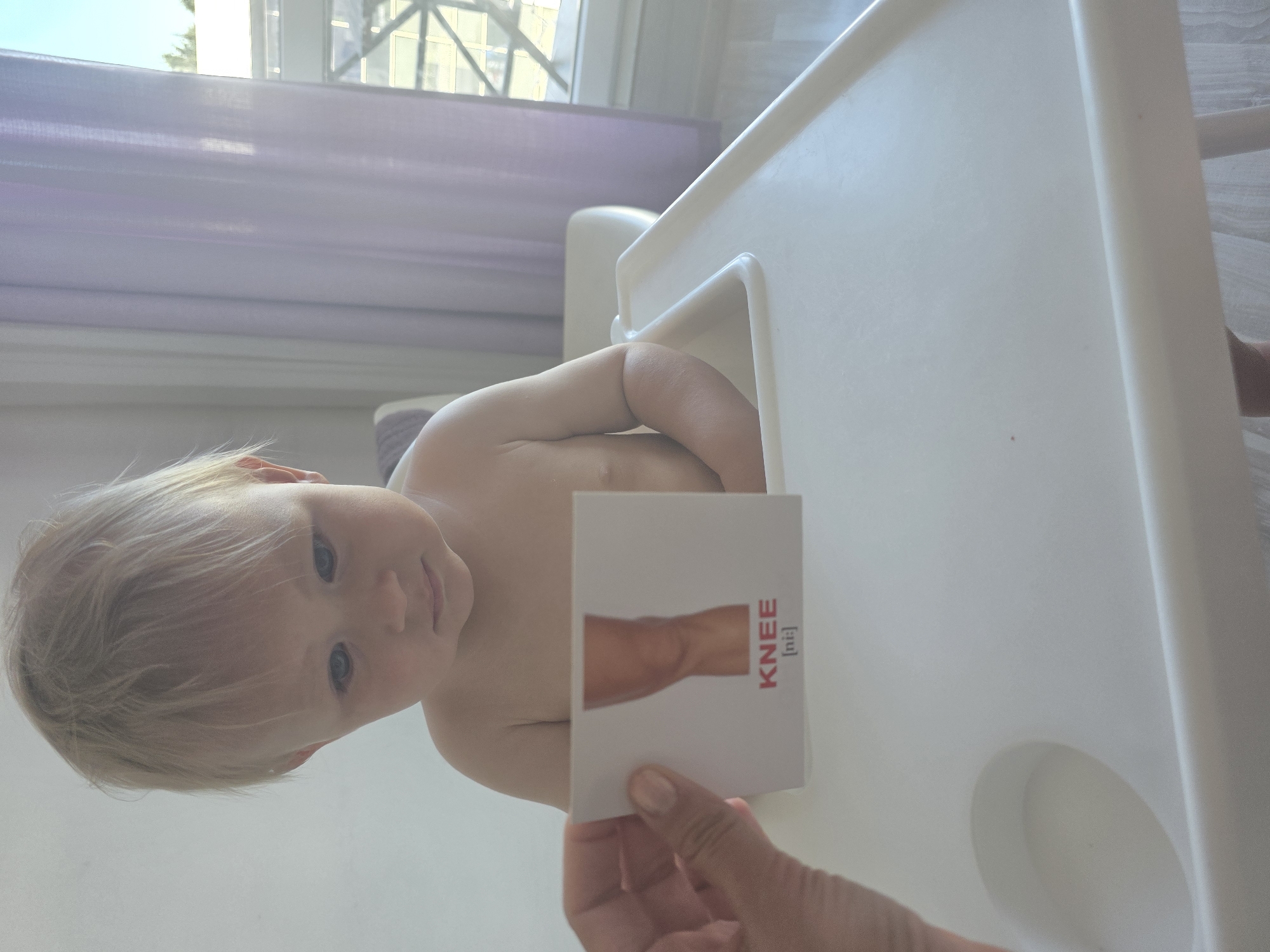 A toddler with blonde hair sits in a white high chair. The toddler is looking down at a flashcard held by an unseen person. The flashcard shows a picture of a knee and the word "KNEE" written below it. The background shows a window with light purple curtains and a glimpse of trees outside. The floor is light-colored wood. The location is a home environment.