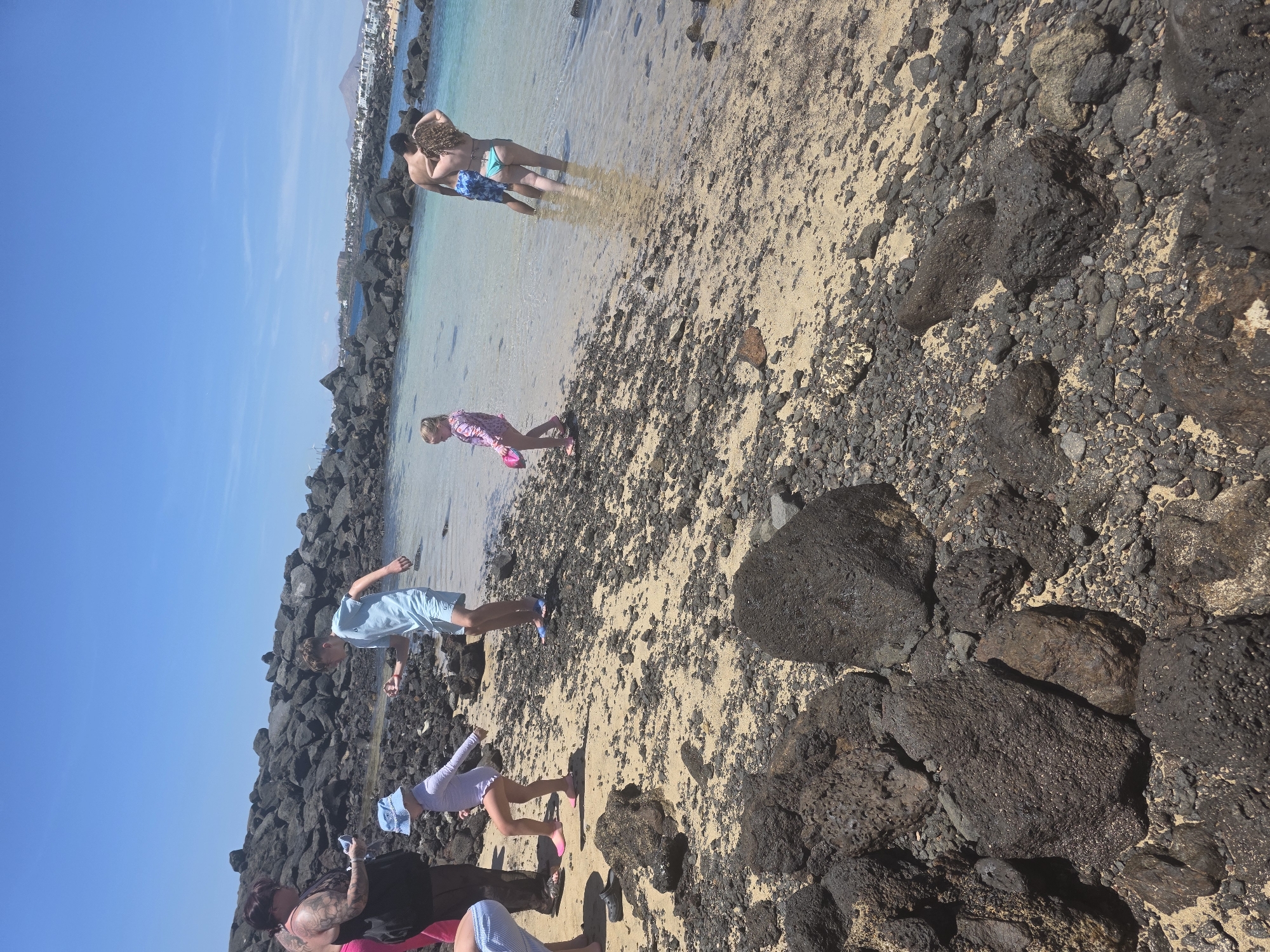 In this daytime, sunny outdoor scene on a sandy beach with dark volcanic rocks, a group of people are enjoying themselves by the water. A couple, partially submerged in the clear, turquoise water, appear to be embracing or interacting intimately. Nearby, a young girl in a pink patterned outfit and pink sandals is walking on the sand, looking down as if at something on the ground. Further down the beach, a boy in light blue shorts and a light blue t-shirt is bending over, perhaps examining something among the rocks. Several other individuals, including a woman with tattoos and a child wearing a blue hat, are also present on the beach, though their activities are less distinct. The background reveals a calm sea meeting the shore, with a line of dark, rocky structures along the coastline, and buildings visible in the distance. The overall atmosphere suggests a relaxed and pleasant day at the beach, likely in a tourist area given the presence of the buildings. No visible text is present in the image.