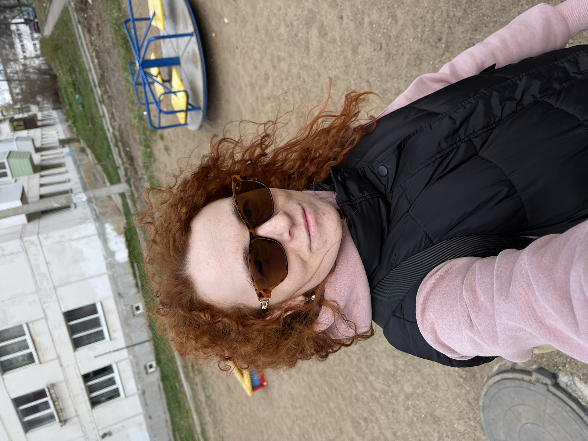 The image is a selfie taken outdoors in what appears to be a playground area, near an apartment building. The person taking the picture is a woman with red curly hair and sunglasses. She is wearing a pink sweatshirt and a black puffy vest. In the background, there's a blue and yellow carousel. Also visible are a sandy ground, a building with white facade, and green patches of grass. The location is near residential buildings, potentially in the city of Chaikovsky, Russia.