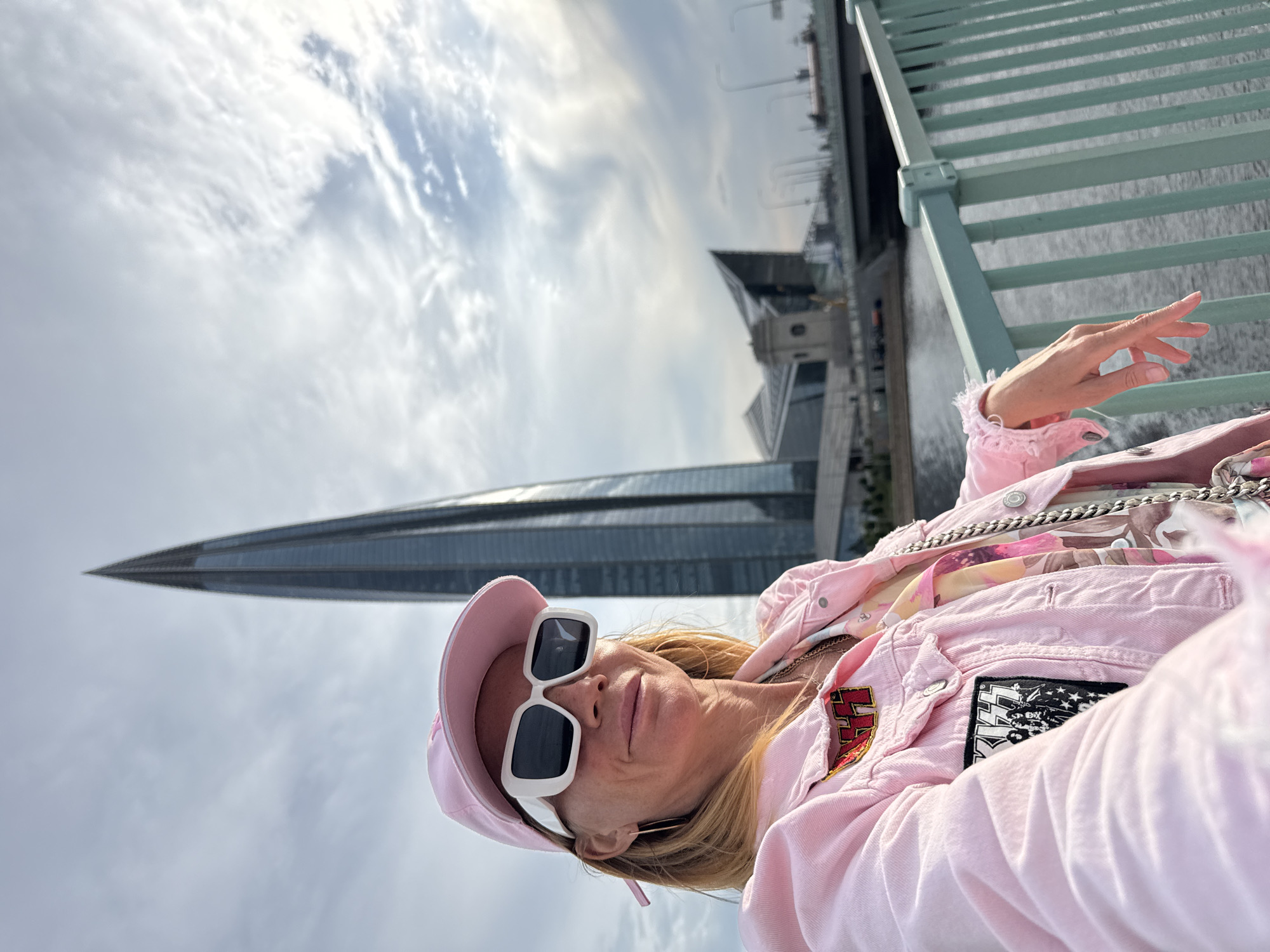 The image, taken in Saint Petersburg, Russia, features a woman in the foreground posing for a selfie. She is wearing a pink baseball cap, large white-framed sunglasses, and a pink denim jacket with patches. Underneath the jacket, she wears a colorful floral-patterned garment. Her left hand is visible, extended towards the camera with painted fingernails.
In the background, a distinct triangular skyscraper, likely the Lakhta Center, dominates the scene. Beyond the skyscraper, a bridge with green railings is visible, and further in the distance, there are other buildings and structures indicative of a cityscape. The sky is filled with dramatic clouds, suggesting either early morning or late afternoon. The overall mood appears cheerful and confident, with the woman smiling at the camera. The lighting suggests it is daytime.