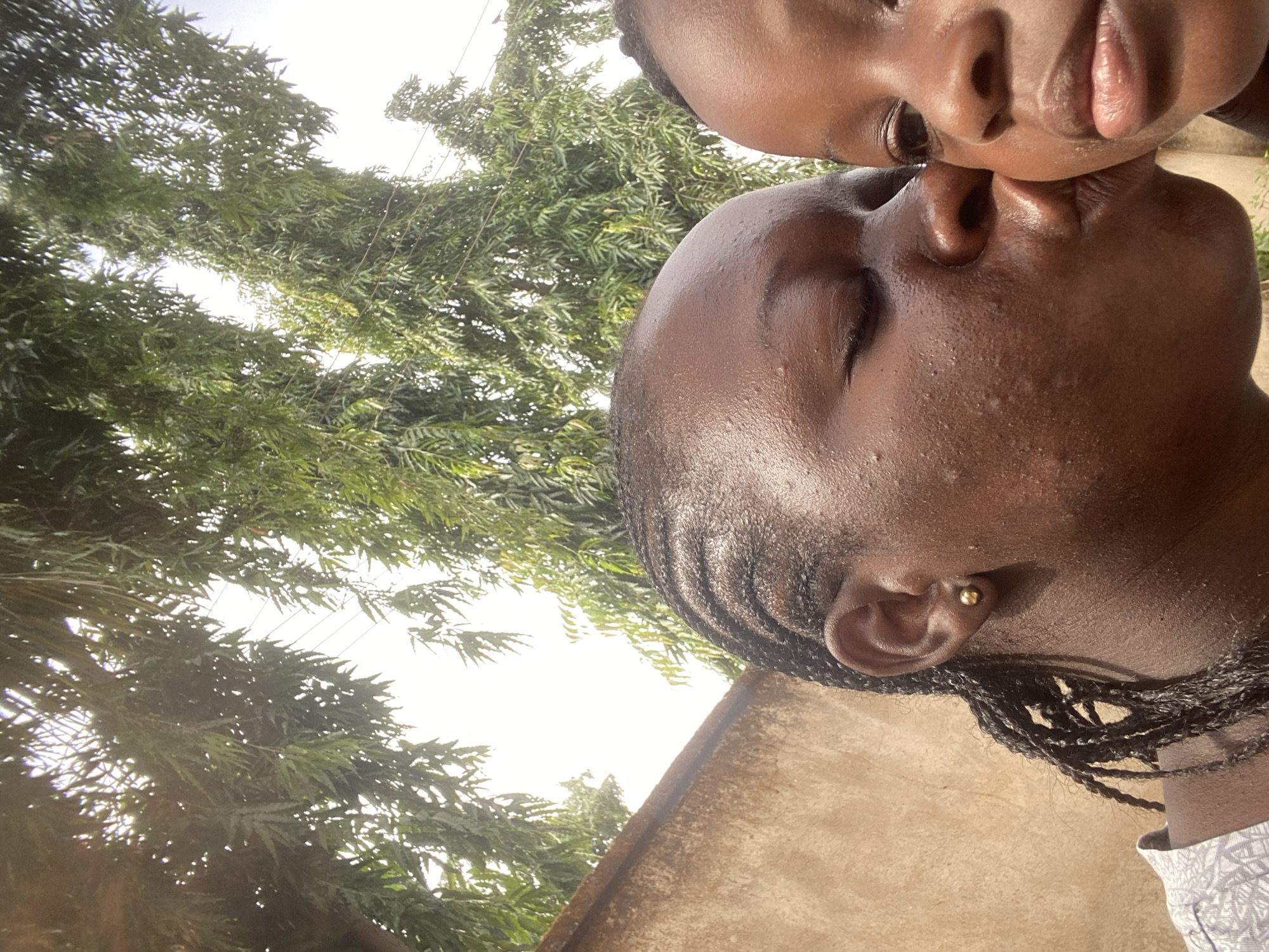 Two dark-skinned individuals are closely positioned with their noses touching in an outdoor setting. The lower individual has their eyes closed and their hair is styled in braids, adorned with a gold stud earring. The person above has their eyes open. Lush, green trees are in the background, filling the upper left quadrant of the image. The location appears to be near a building with a light-colored exterior. The location is Maigizo, Nigeria.
