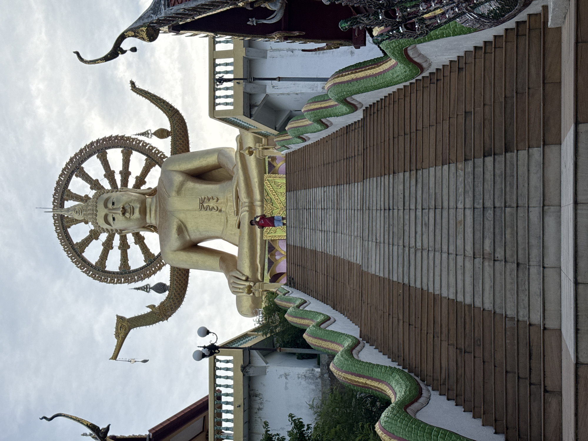 The image shows a large golden Buddha statue in Baan Plai Laem, Thailand. The statue is seated with a wheel of dharma behind its head and is positioned at the top of a long, wide staircase. The staircase has a decorative railing on either side, shaped like a colorful serpent or naga. At the bottom of the staircase, the ground is paved. In the middle of the staircase, a person wearing a red shirt and blue shorts is standing and taking a photo with a mobile device. The sky is overcast. The architecture surrounding the statue and staircase appears to be a Buddhist temple complex, with ornate roofs and decorative elements visible.