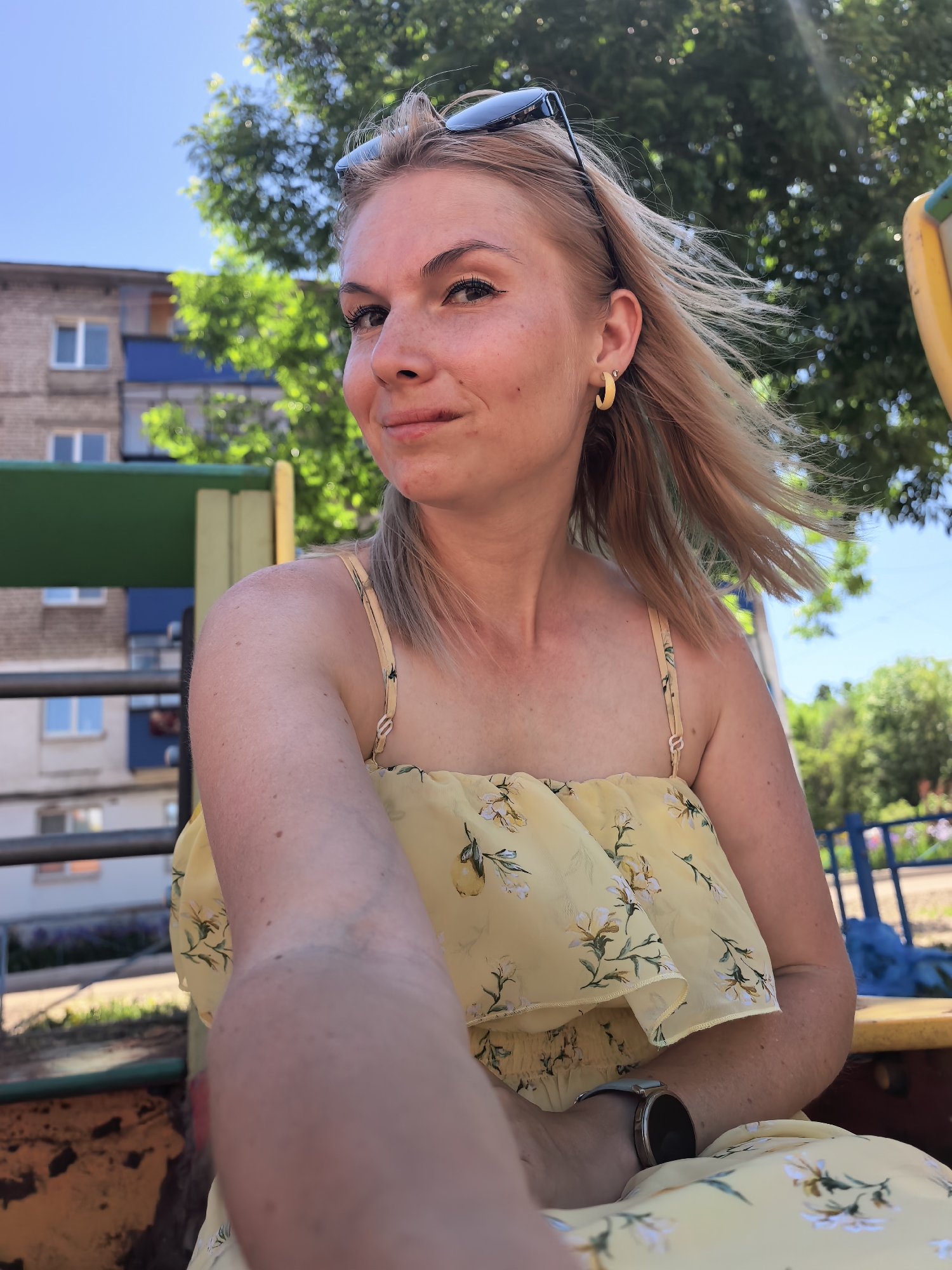 A woman poses for a selfie outdoors. She has fair skin and blonde hair, and she's wearing a yellow floral dress. Sunglasses are perched on her head. A gold hoop earring is visible in her left ear. She is on a playground and appears to be sitting on play equipment. In the background is a residential building, trees, and blue sky. This playground is located in Sterlitamak, Russia.