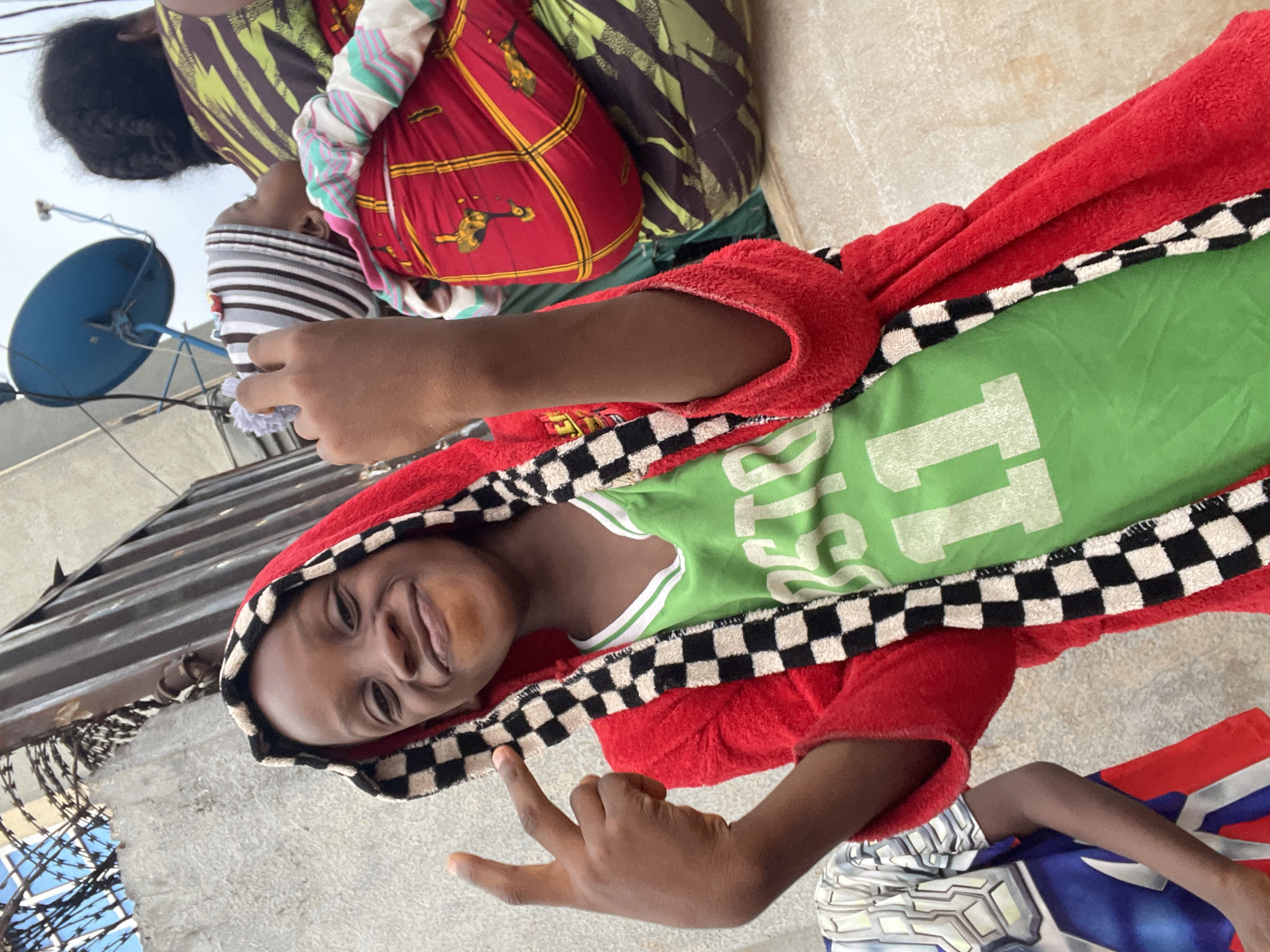 A young person with dark skin, wearing a green t-shirt with white lettering and a red hooded blanket with a black and white checkered trim, is smiling and gesturing with their hand. They are located on what appears to be a rooftop in Onitsha, Nigeria, as indicated by the presence of a satellite dish, barbed wire fencing, and corrugated metal roofing. The scene includes additional individuals in the background, wrapped in colorful fabrics. The setting suggests an urban residential area.