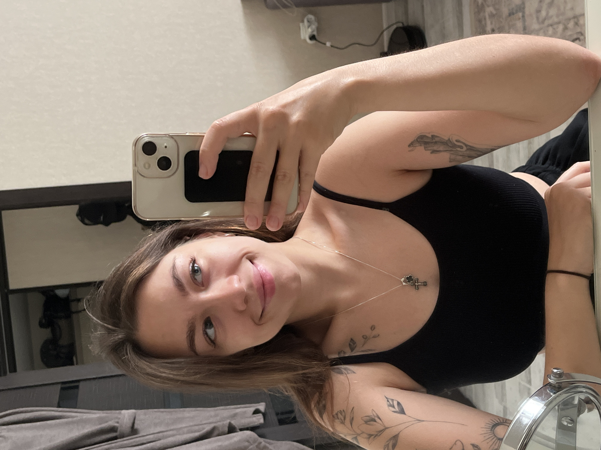 A young woman with long brown hair, wearing a black tank top and a silver necklace with a cross pendant, is taking a mirror selfie. She has tattoos on her left arm and shoulder. The background appears to be an indoor setting, possibly a bedroom, with a closet, a mirror, and a wall with an electrical outlet. The lighting is natural and comes from the right. The woman is smiling and looking towards the mirror. The image is taken at a slight angle, showing her face and upper body. Based on the provided image, it is impossible to determine the specific location within the city of Chaikovsky, Russia.
