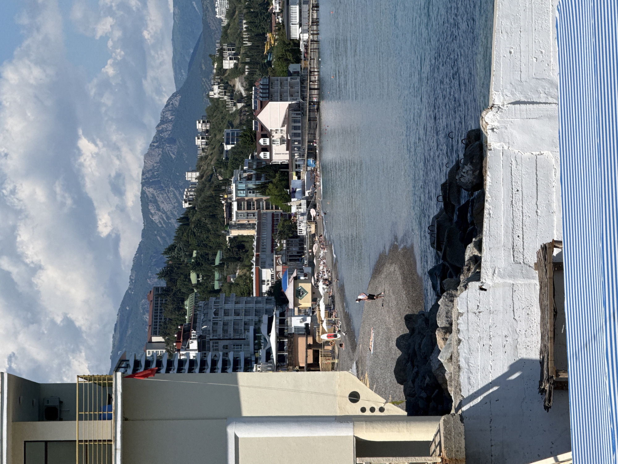 A vibrant coastal landscape is captured from an elevated viewpoint, likely a balcony, on a bright, partly cloudy day. In the immediate foreground, a sturdy, weathered white concrete structure partially frames the scene, with a fragment of blue and white striped fabric (possibly an awning or screen) visible on the far right.
Beyond this structure, the calm, blue-green sea extends towards the horizon, gently rippling under the sunlight. A pebbly beach runs along the shoreline, dotted with several people enjoying the day. One individual is seen walking near the water's edge, while another lies on the sand, presumably sunbathing. Beach umbrellas and loungers are scattered across the beach, suggesting a resort or recreational area. A dark, rocky breakwater or natural rock formation separates the foreground from the main beach.
Along the coast, a bustling town or resort is densely built, with buildings stretching along the beachfront and ascending the lower slopes of the mountains. A variety of architectural styles are present, many featuring balconies and flat roofs, nestled among lush green vegetation and trees.
In the background, grand, rugged mountains rise steeply, showcasing both forested areas and exposed rock faces. The sky above is a clear blue, adorned with scattered white cumulus clouds. The scene depicts a sunny day, likely mid-morning to late afternoon, conveying a peaceful, leisurely atmosphere. No discernible text is visible anywhere in the image.